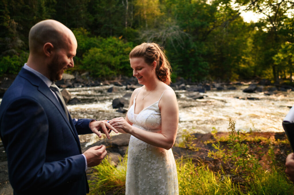 A couple stands by a riverside; the woman is placing a ring on the man's finger. They are in a forest setting, with sunlight filtering through the trees. The man is wearing a suit, and the woman is in a white dress, both smiling warmly.