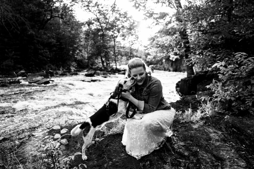 A woman kneels by a river, hugging a small dog. She wears a long dress and a denim jacket. The scene is surrounded by trees and foliage, with sunlight filtering through. It's a serene, black-and-white nature setting.
