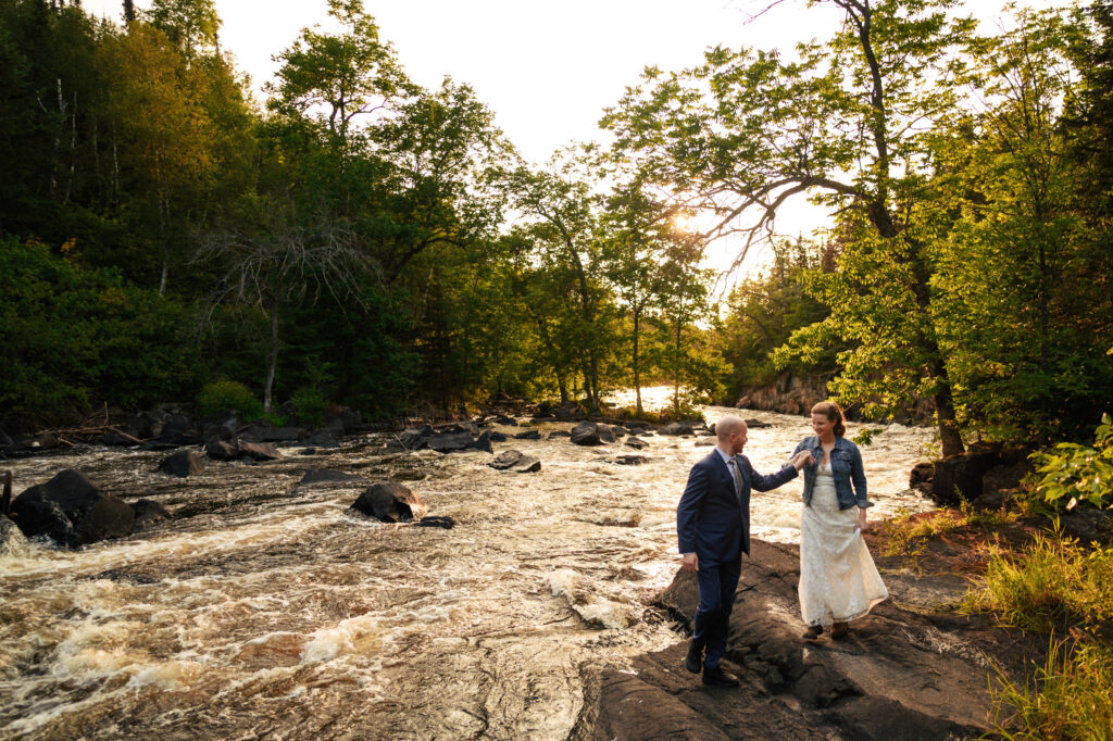 A couple stands by a flowing river surrounded by lush greenery. The man, in a suit, holds the hand of the woman in a white dress and denim jacket as they walk along the rocky riverbank under a bright, sunlit sky.