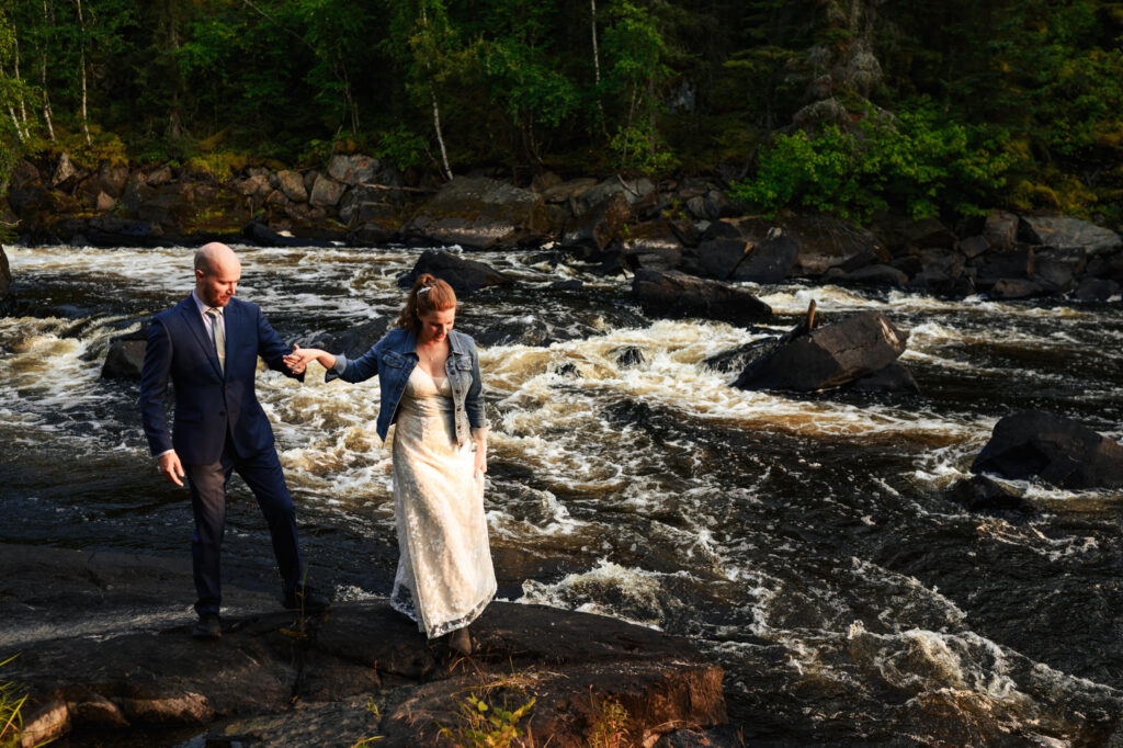 A couple is walking on rocks by a flowing river. The man is wearing a suit, and the woman is in a white dress and denim jacket. They are surrounded by greenery and trees, and the sun casts a warm glow on the scene.