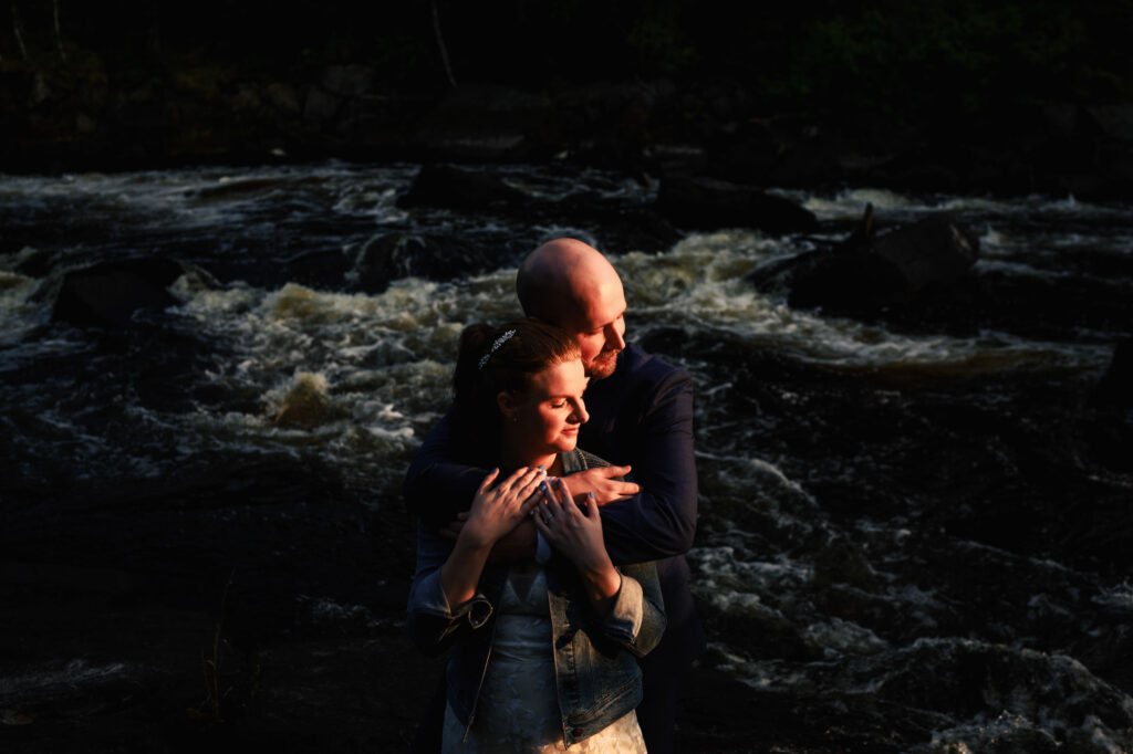 A couple embraces warmly in front of a flowing river. The person in front, wearing a denim jacket, leans back with closed eyes into the arms of the other person, who is smiling and wearing a dark sweater. The scene is dimly lit, highlighting their expressions.