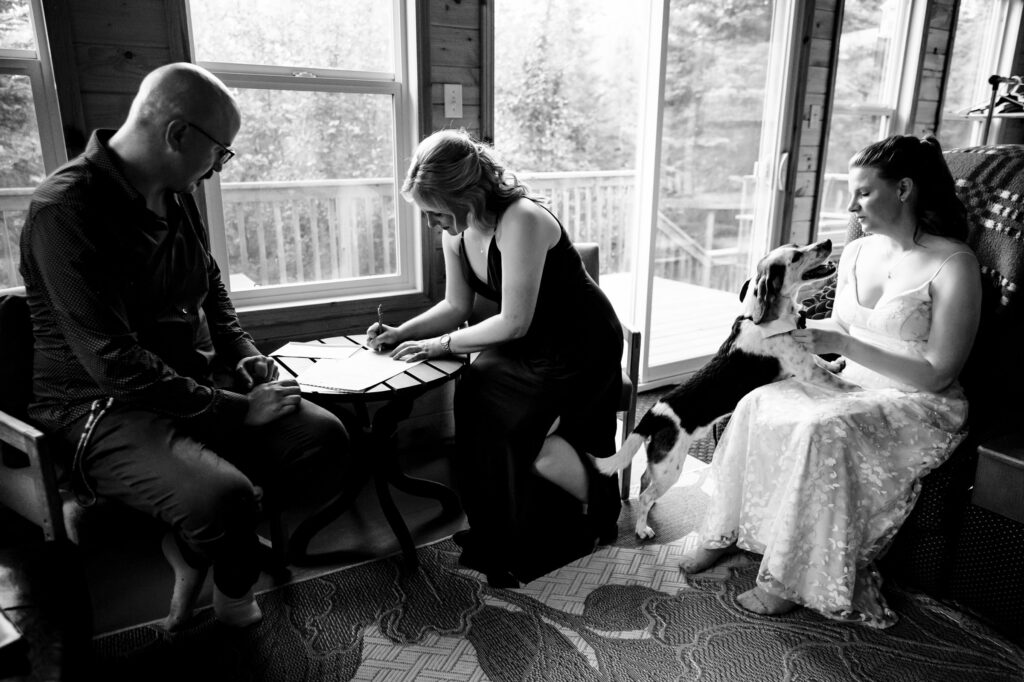 A black and white photo of three people inside a room. A person on the left sits at a table, while another signs a document. A woman in a dress sits on the right, holding a dog. Large windows reveal a wooded view outside.