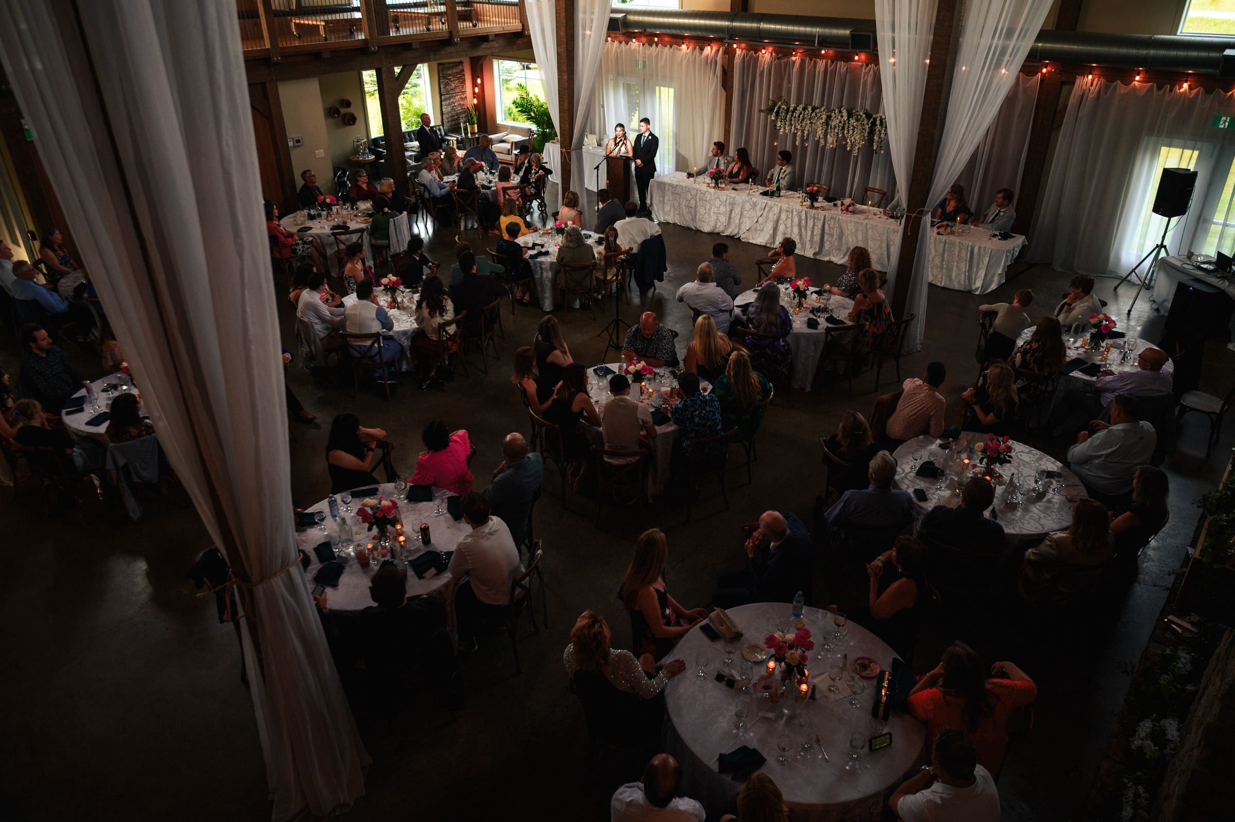 Wedding reception in a dimly lit Winnipeg venue, guests at round tables, head table on stage.