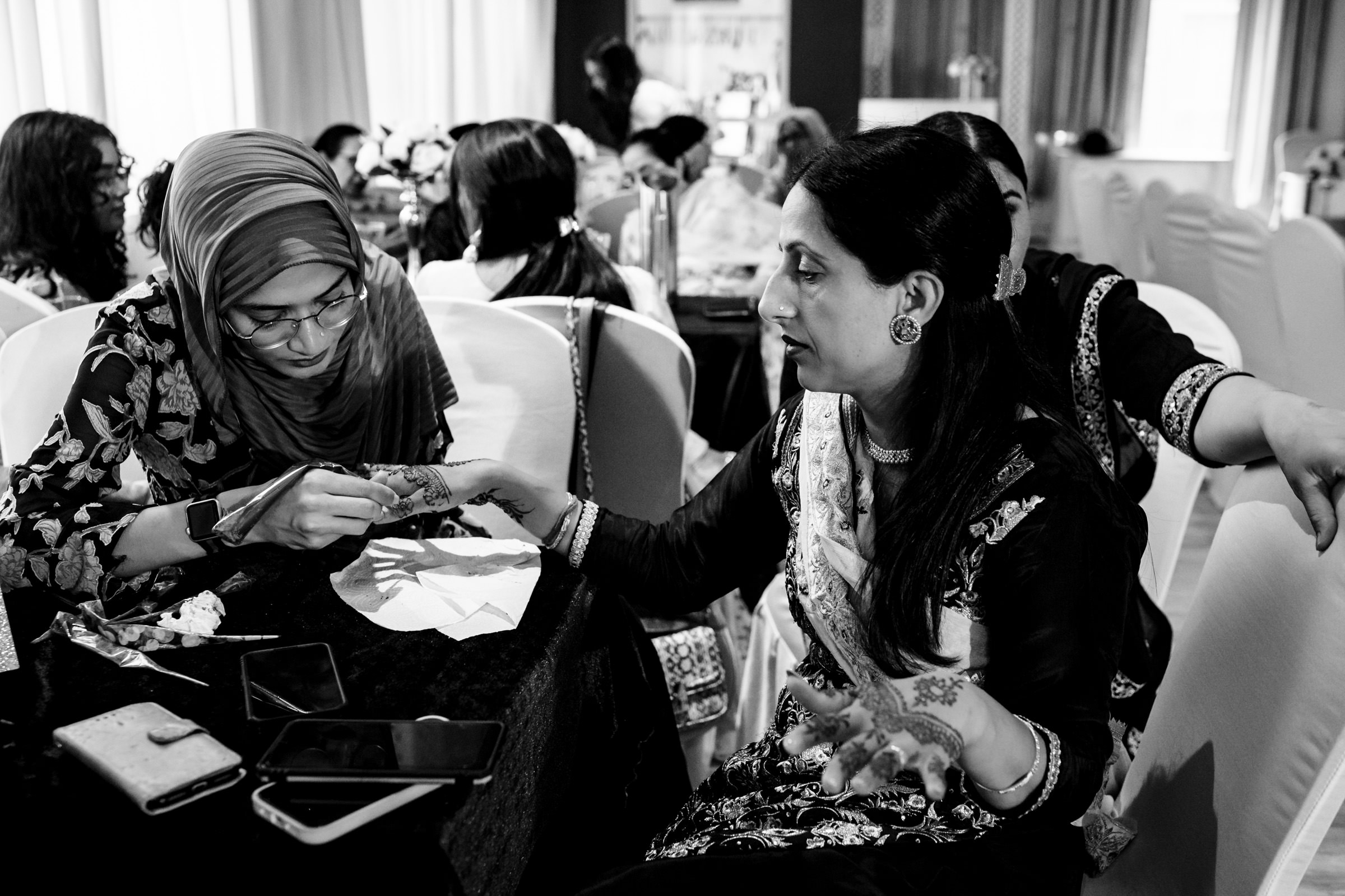 A black and white photo of two women at a gathering. One woman is applying henna on the other's hand. They are seated at a table with smartphones and paper. Several other people are visible in the background, creating a busy atmosphere.