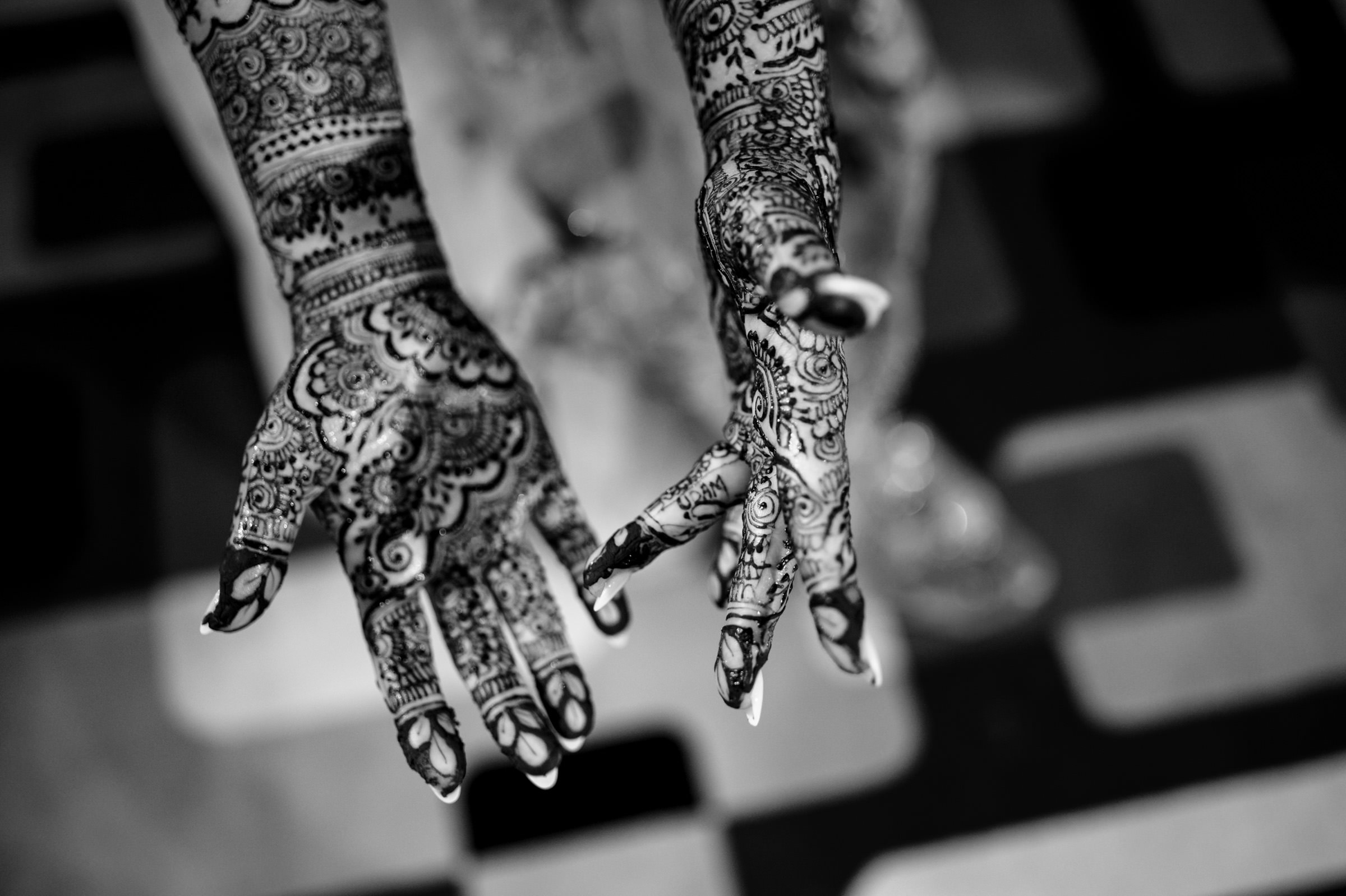 Black and white image of two hands adorned with intricate henna designs. The patterns cover the palms and extend to the fingers, featuring elaborate floral and geometric motifs. The background is a blurred checkerboard pattern.