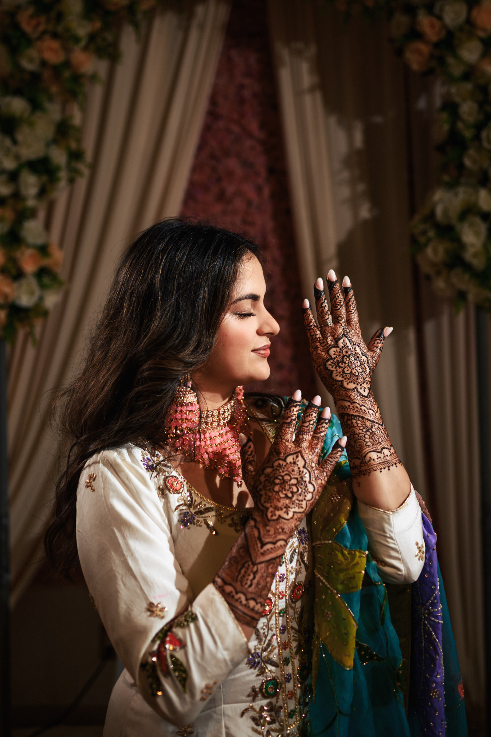A woman with long dark hair, wearing a white and green traditional outfit, intricately decorated with henna on her hands, and adorned with pink floral earrings, stands elegantly with eyes gently closed. Floral arrangements are visible in the background.