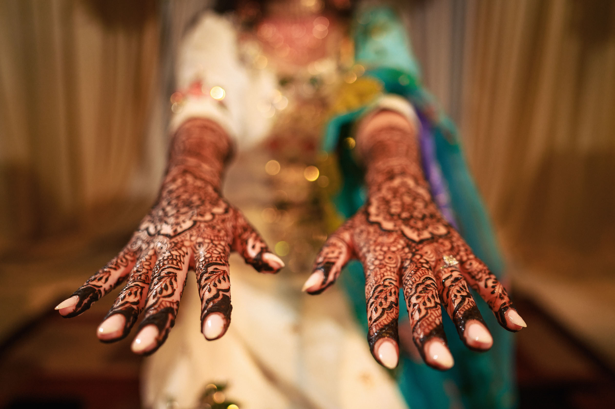 A person holding their hands out, showcasing intricate henna designs. The patterns cover the palms and extend to the fingers. The person is dressed in colorful, ornate fabric, and the background is softly blurred.