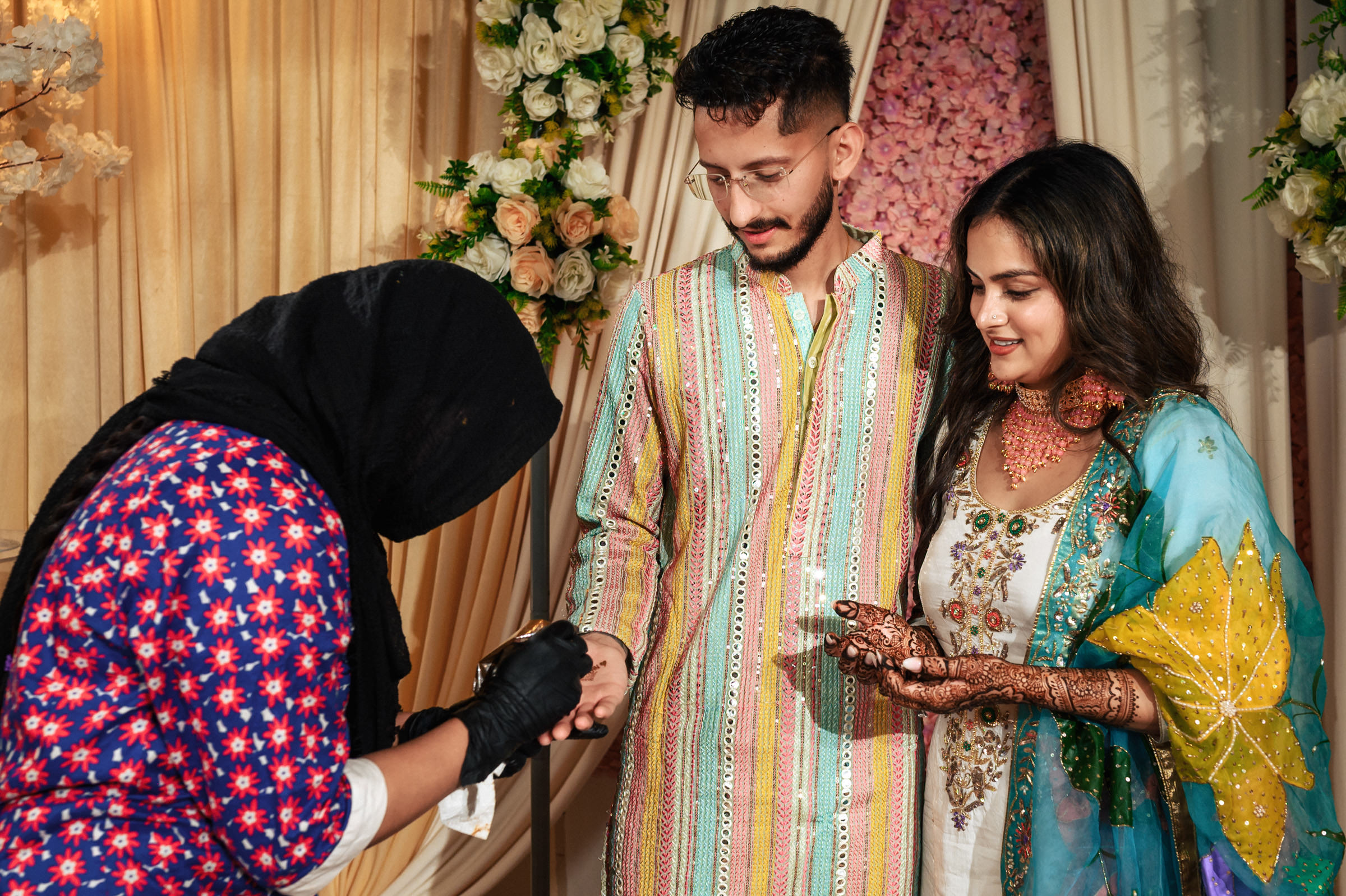 A man and woman in colorful traditional attire participate in a henna ceremony. The woman extends her hand to a henna artist applying intricate designs. The background features floral decorations in a warm, inviting setting.