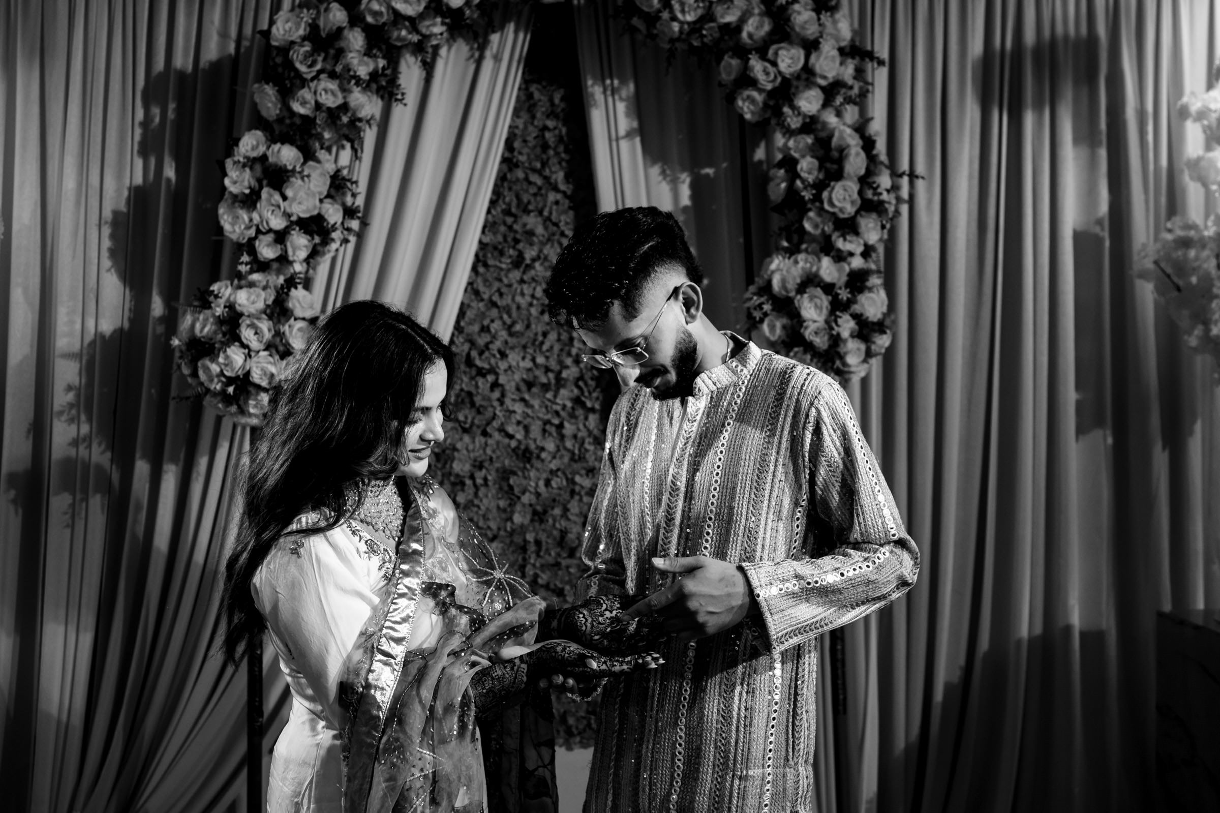 A couple in traditional attire stands together in front of a floral backdrop. The woman is holding a decorative item, while both look at it. The scene is lit softly, creating a warm and intimate atmosphere.