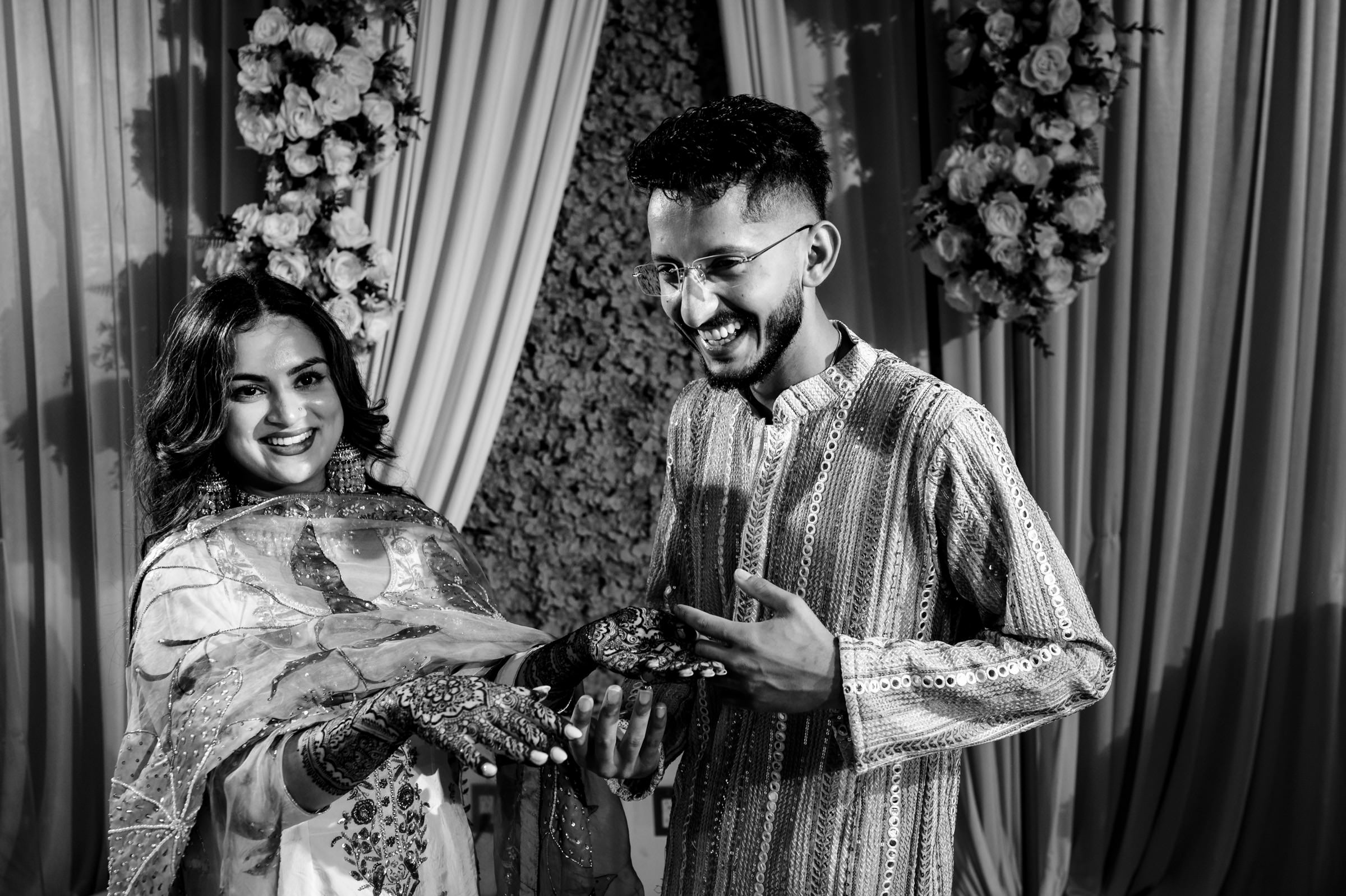 A smiling couple celebrates an engagement. The woman displays intricate henna designs on her hands. They stand in front of draped curtains adorned with floral arrangements.