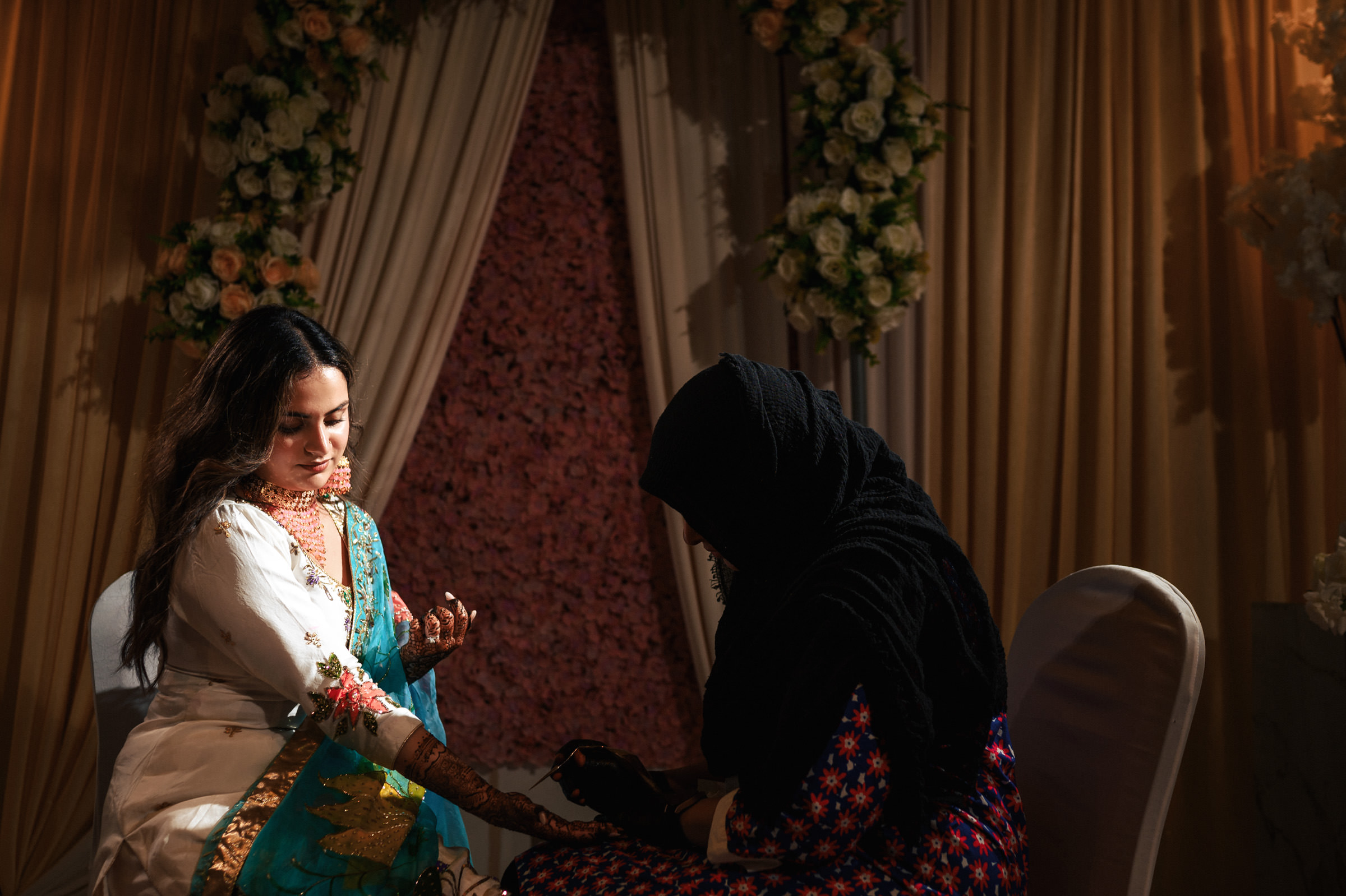 A woman in a white and teal dress is getting intricate henna designs applied to her hand by another woman in a dark outfit. They are seated indoors, surrounded by floral decorations and draped curtains, under warm lighting.