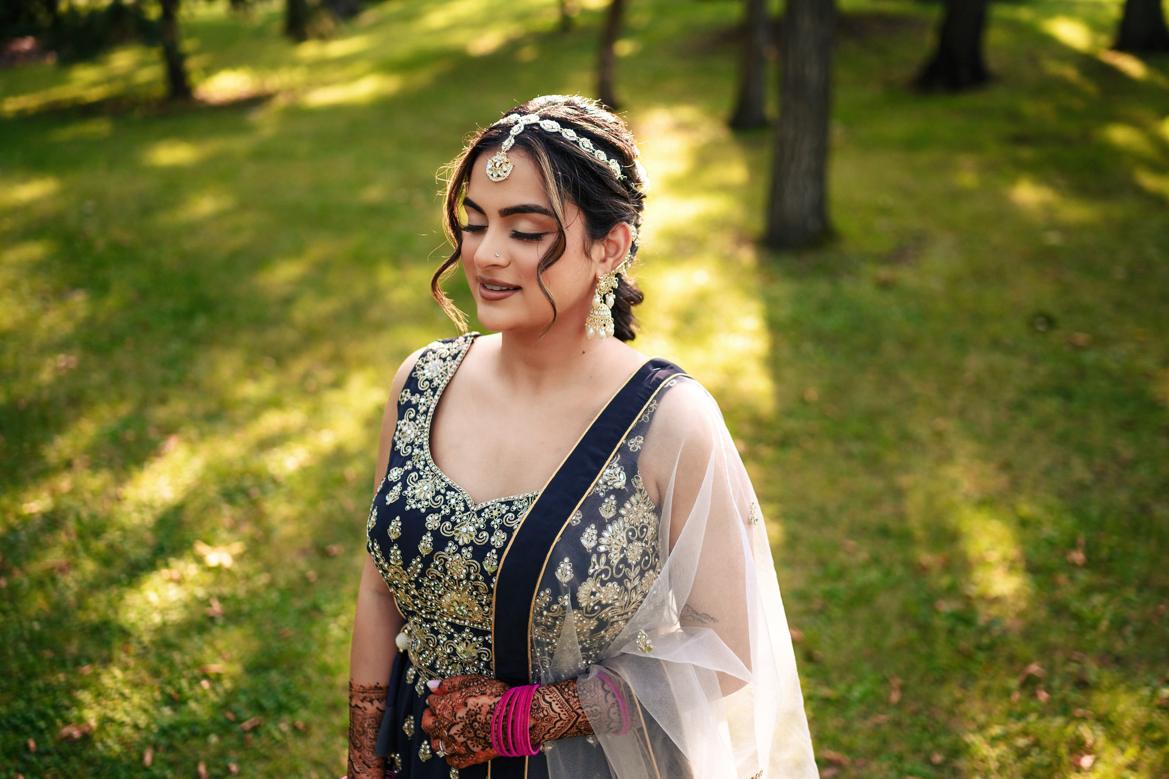 A woman stands outdoors on grass, wearing an ornate blue and gold traditional outfit. Her hair is styled with decorative hairpieces, and she has henna on her hands. Sunlight filters through the trees, creating a dappled light effect around her.