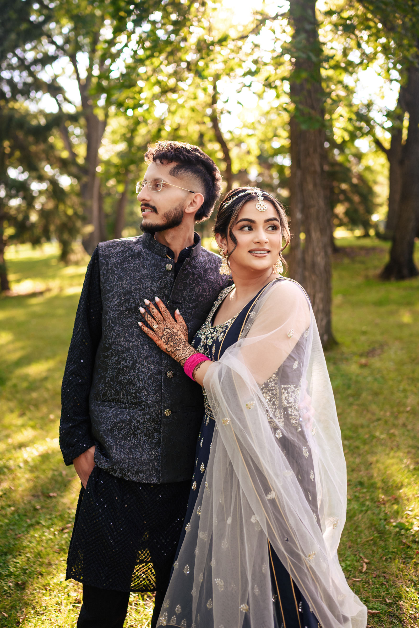 A couple dressed in traditional attire stands outdoors in a sunlit forest area. The woman wears a decorative outfit with henna on her hands, while the man is in a dark ethnic suit. Both look to the side, surrounded by green trees and grass.