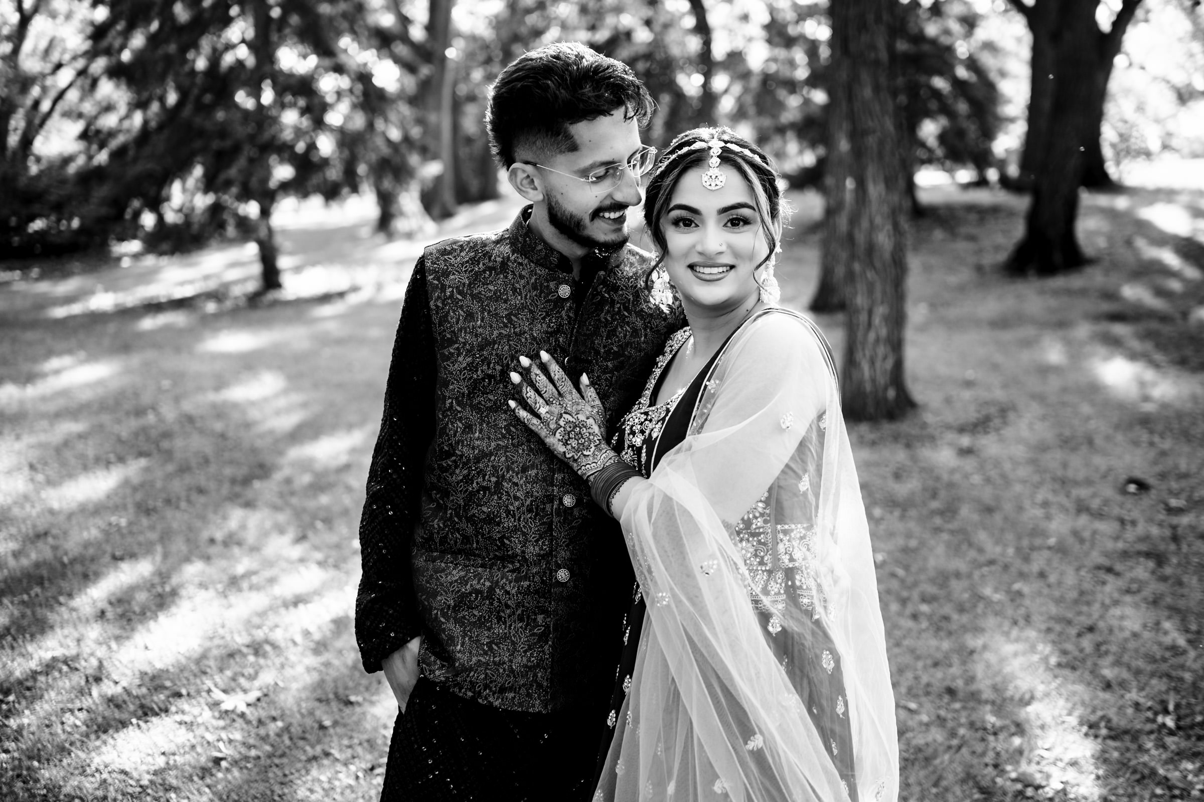 A couple poses in a sunlit park, wearing traditional South Asian attire. The woman, adorned with intricate jewelry and henna, embraces the man as they both smile warmly, surrounded by trees and dappled sunlight.