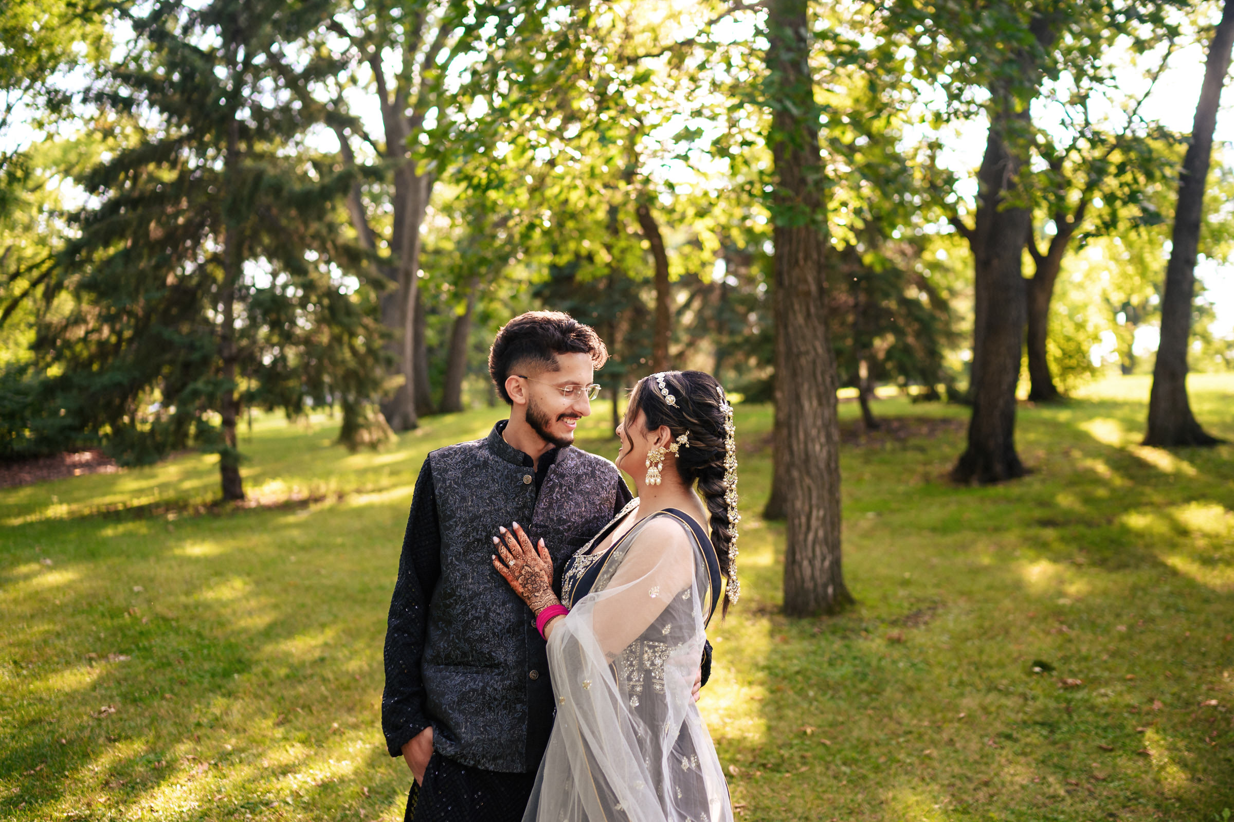 A couple stands together in a sunlit park, surrounded by trees. They are smiling at each other, with the woman wearing traditional attire and the man in a dark jacket. The grass and trees create a serene background.
