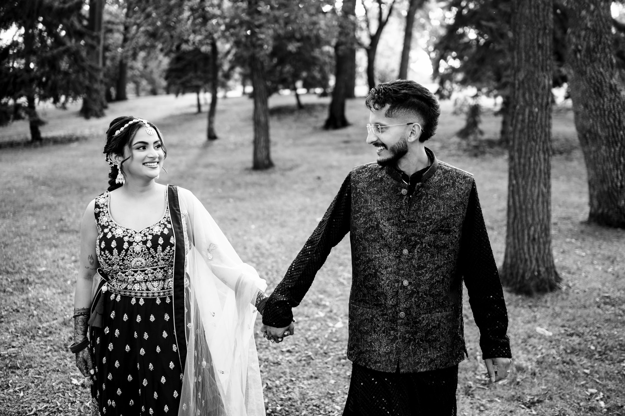 A couple dressed in traditional attire walk hand in hand through a wooded park. Both are smiling and looking at each other. The image is in black and white, capturing a joyful moment amidst the trees.