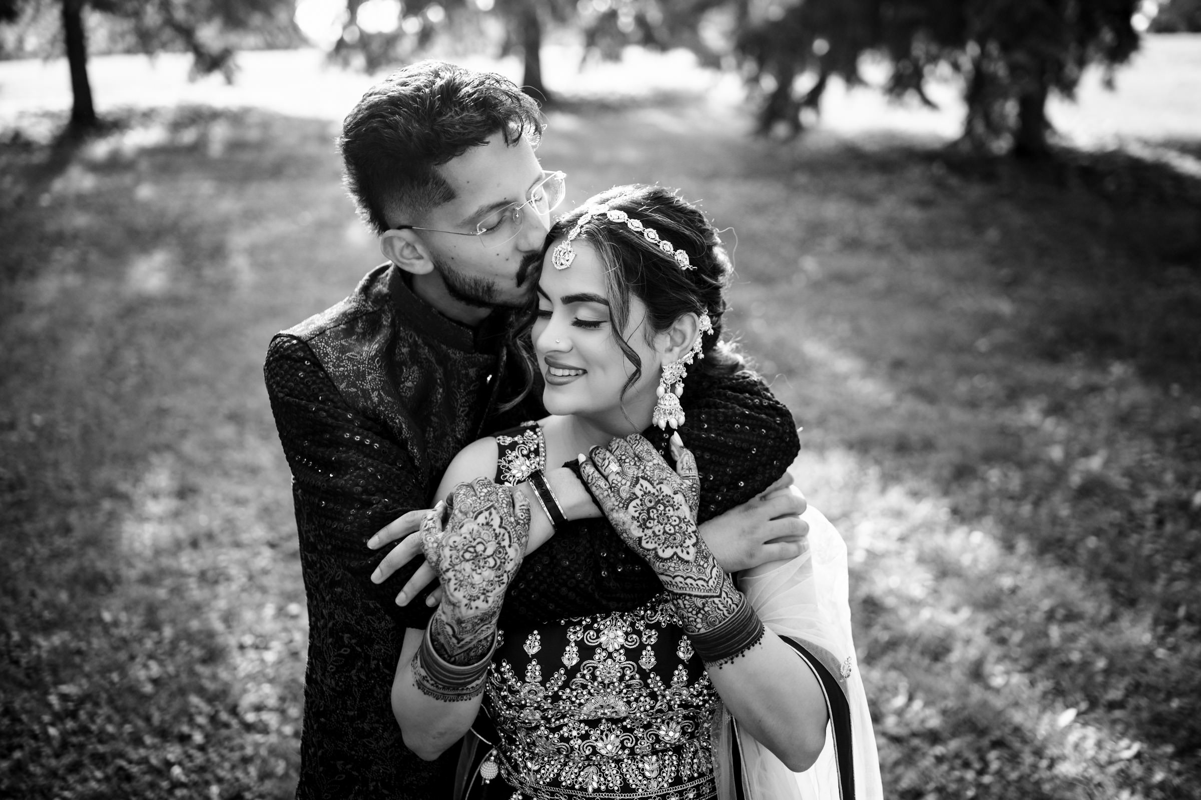 A black and white photo of a couple in traditional attire, standing in a grassy area. The man embraces the woman from behind, kissing her head. She is smiling, her hands adorned with intricate henna designs, bangles, and jewelry.