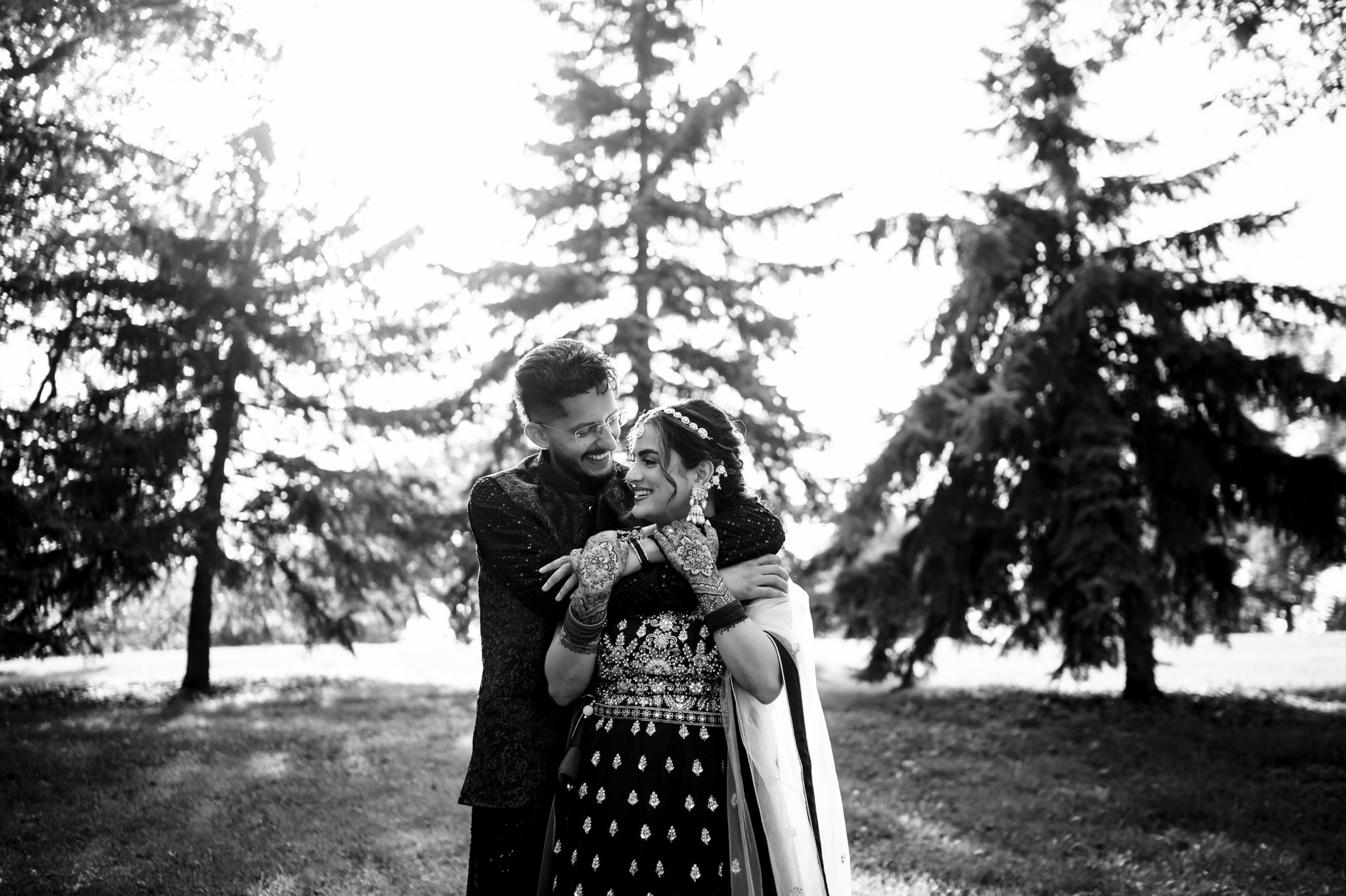 A couple in traditional attire embrace joyfully in a sunlit park with tall trees in the background. The woman's hands are decorated with henna designs, and they are both smiling. The scene captures a moment of happiness and love.