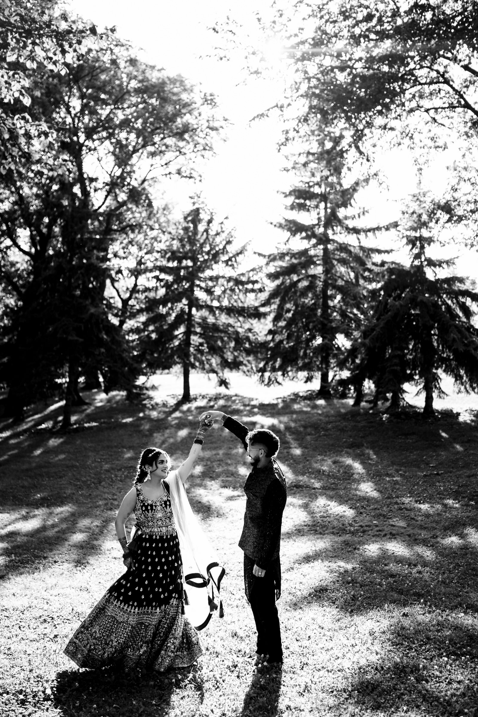 A couple dances joyfully on a grassy lawn surrounded by tall trees. The woman wears a traditional flowing dress and the man is in a suit. They appear carefree, with sunlight filtering through the trees, creating intricate shadows on the ground.