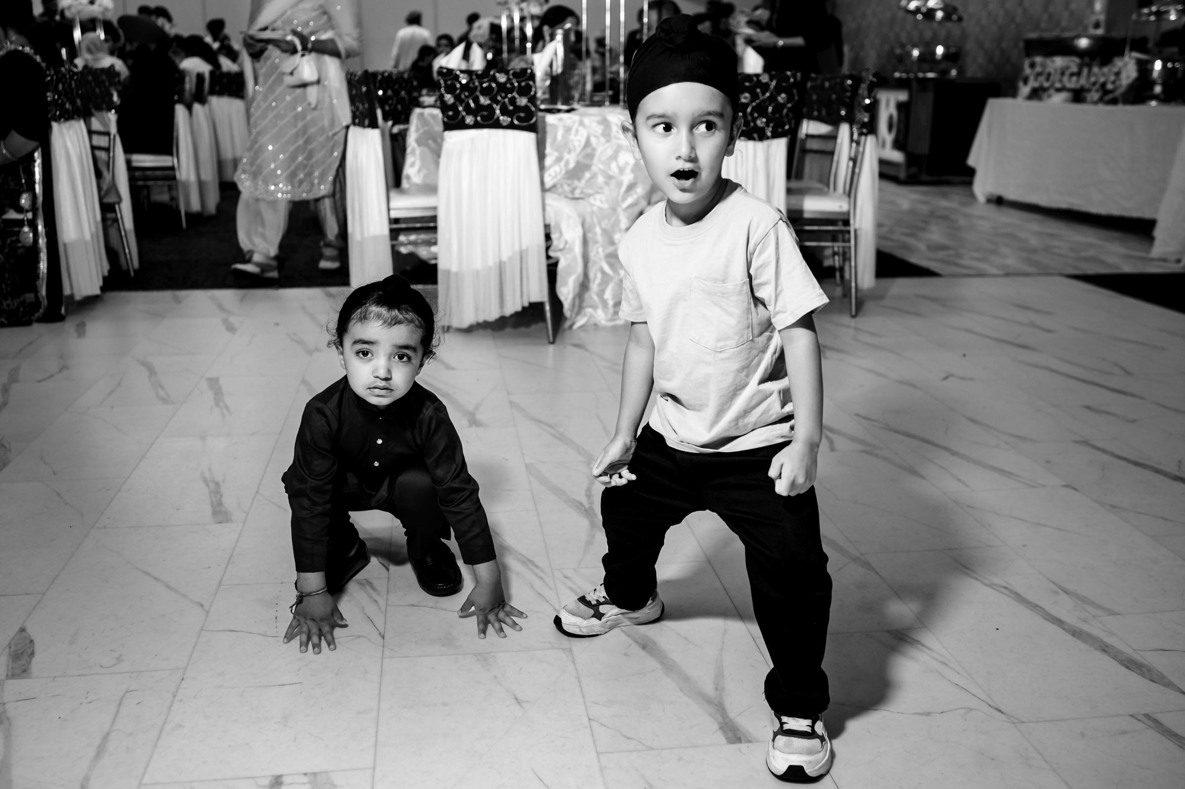 Two young children are playfully posing indoors. One child in a light shirt and dark pants crouches down, while the other, in a dark outfit and head covering, stands with a surprised expression. The background shows a gathering with people and tables.