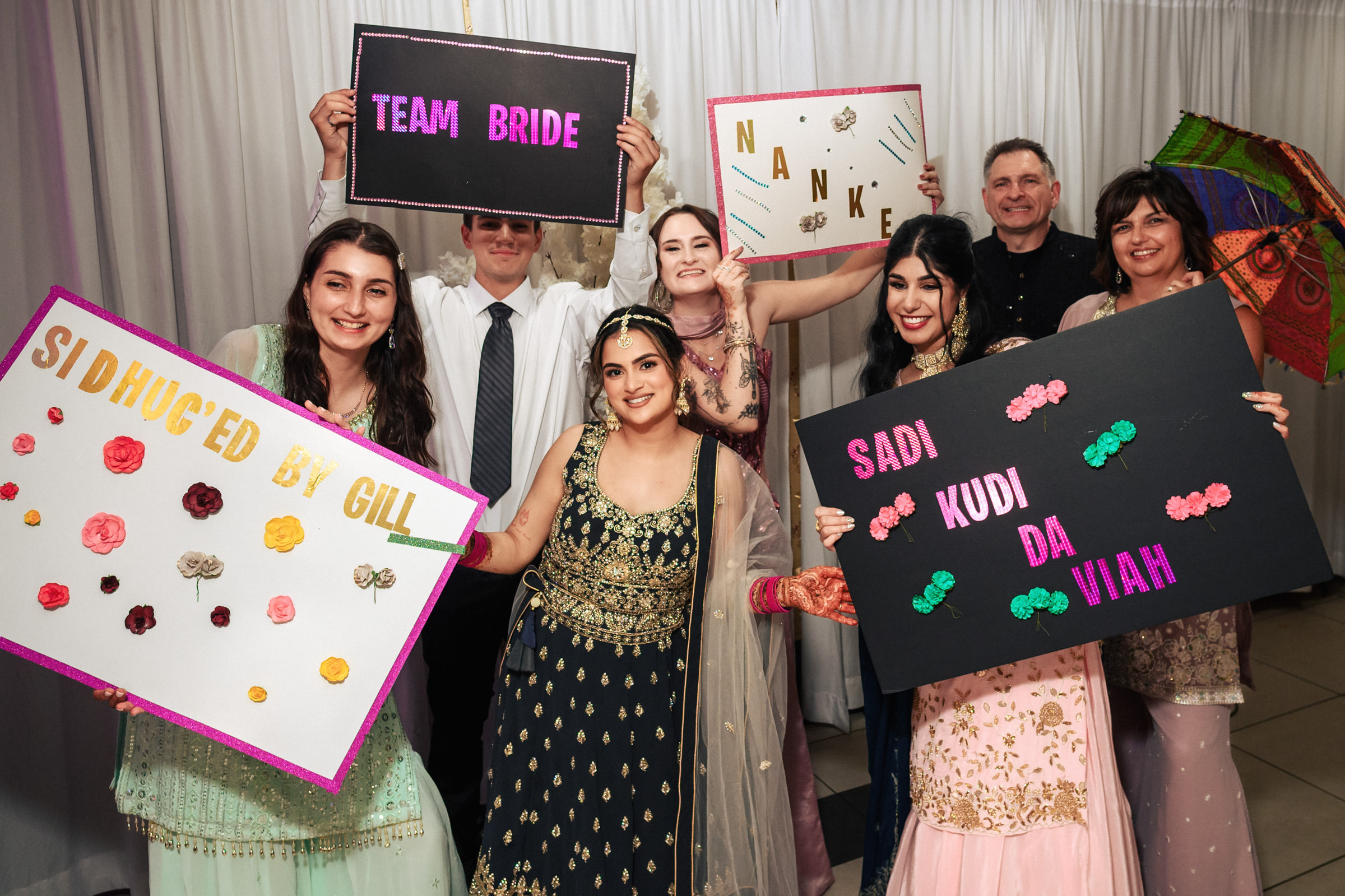 A joyful group of people dressed in festive attire holding colorful signs with celebratory messages, including "Team Bride" and "Sadi Kudi Da Viah." The background features white drapes.
