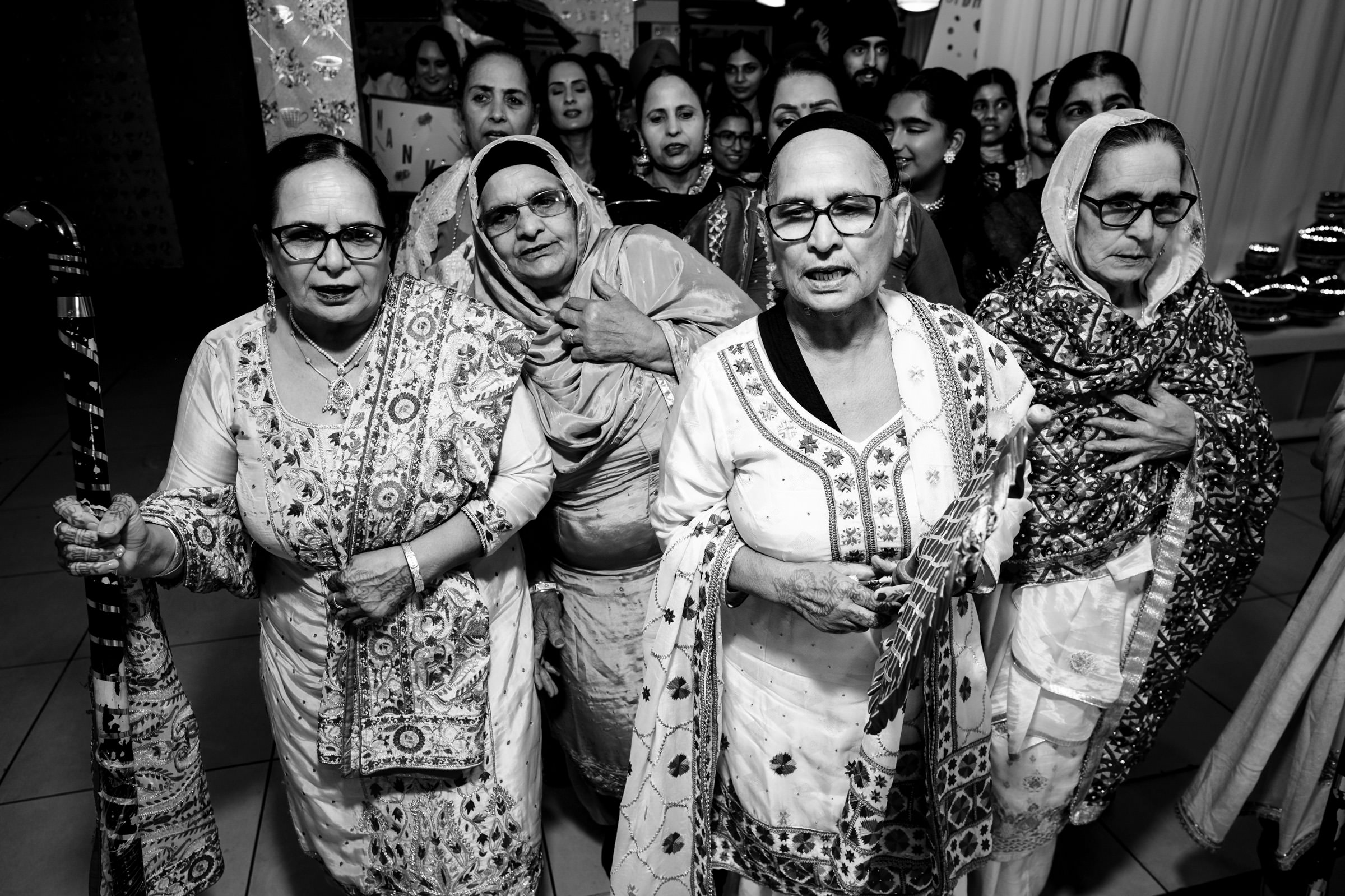 A group of elderly women in traditional attire are gathered closely, expressing joy and camaraderie. They are in a festive indoor setting, wearing patterned shawls and glasses, some holding bamboo sticks. The scene captures a moment of cultural celebration.