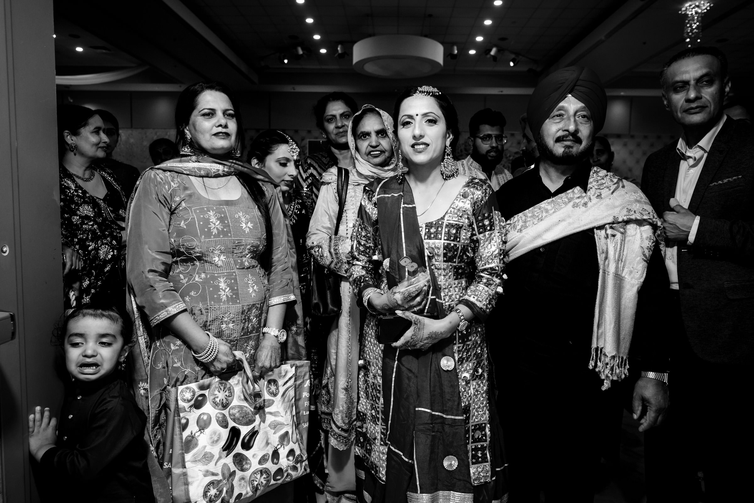 A group of people in traditional attire, including a woman in bridal wear holding a bouquet, gather indoors for a celebration. A young child stands to the side. The atmosphere is joyful and festive. Black and white image.
