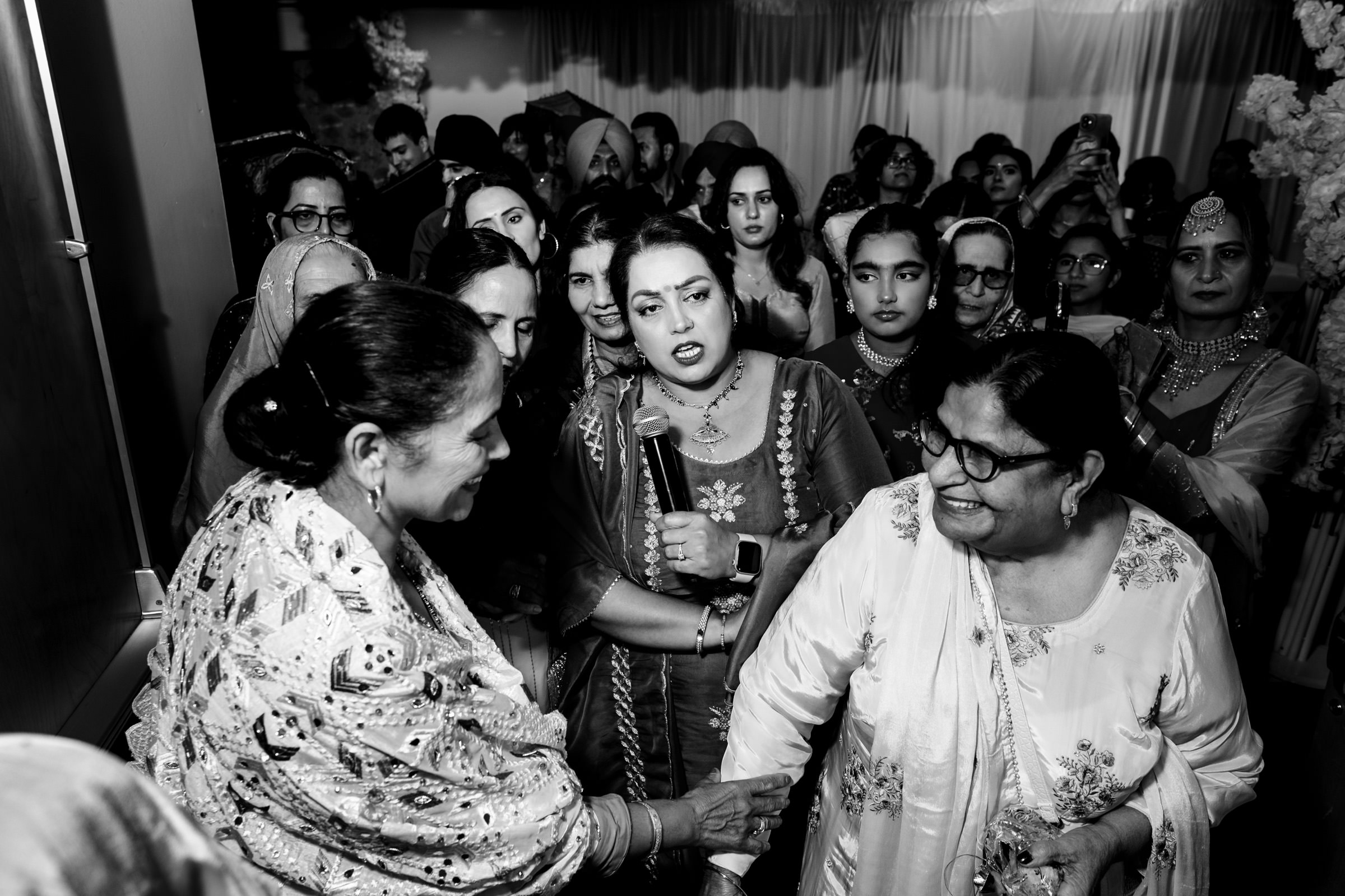 A crowded scene at a wedding ceremony. Women in traditional attire gather closely, with two women interacting in the foreground, one elderly and the other middle-aged. Their expressions show warmth and engagement. Black and white photograph.