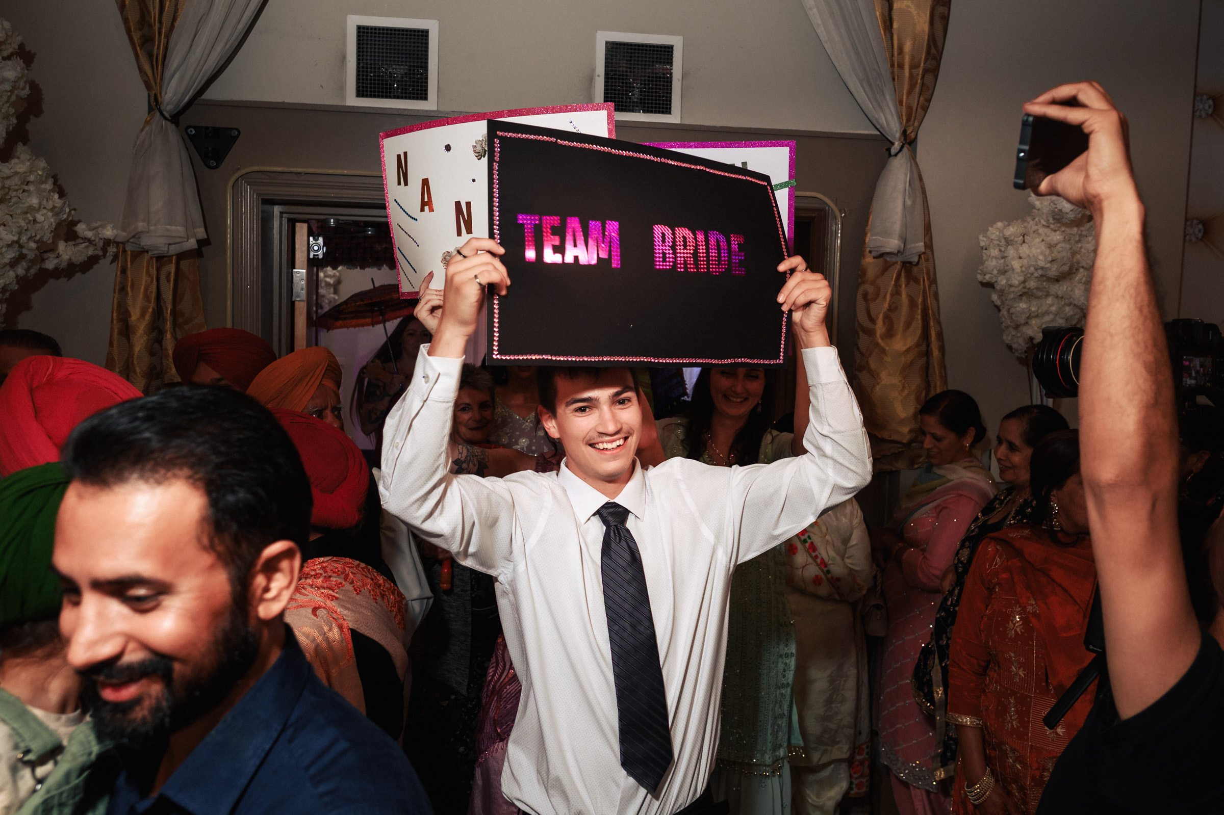 A smiling man in a white shirt and black tie holds up a sign that says "Team Bride" at a lively gathering. People around him are dressed in colorful traditional attire, and the atmosphere is festive.