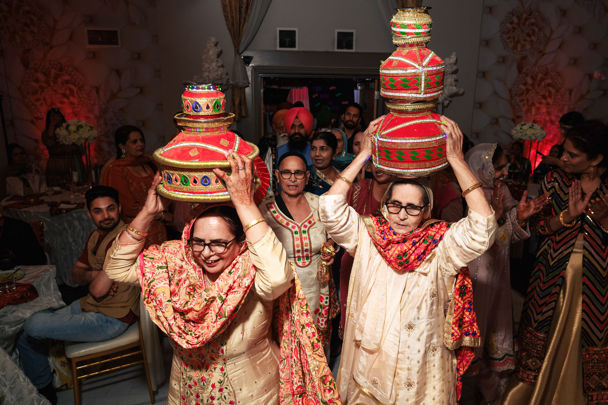 Two women in traditional attire carry colorful, decorated vessels on their heads during a cultural celebration. They are accompanied by a group of people clapping and cheering in a festive indoor setting.