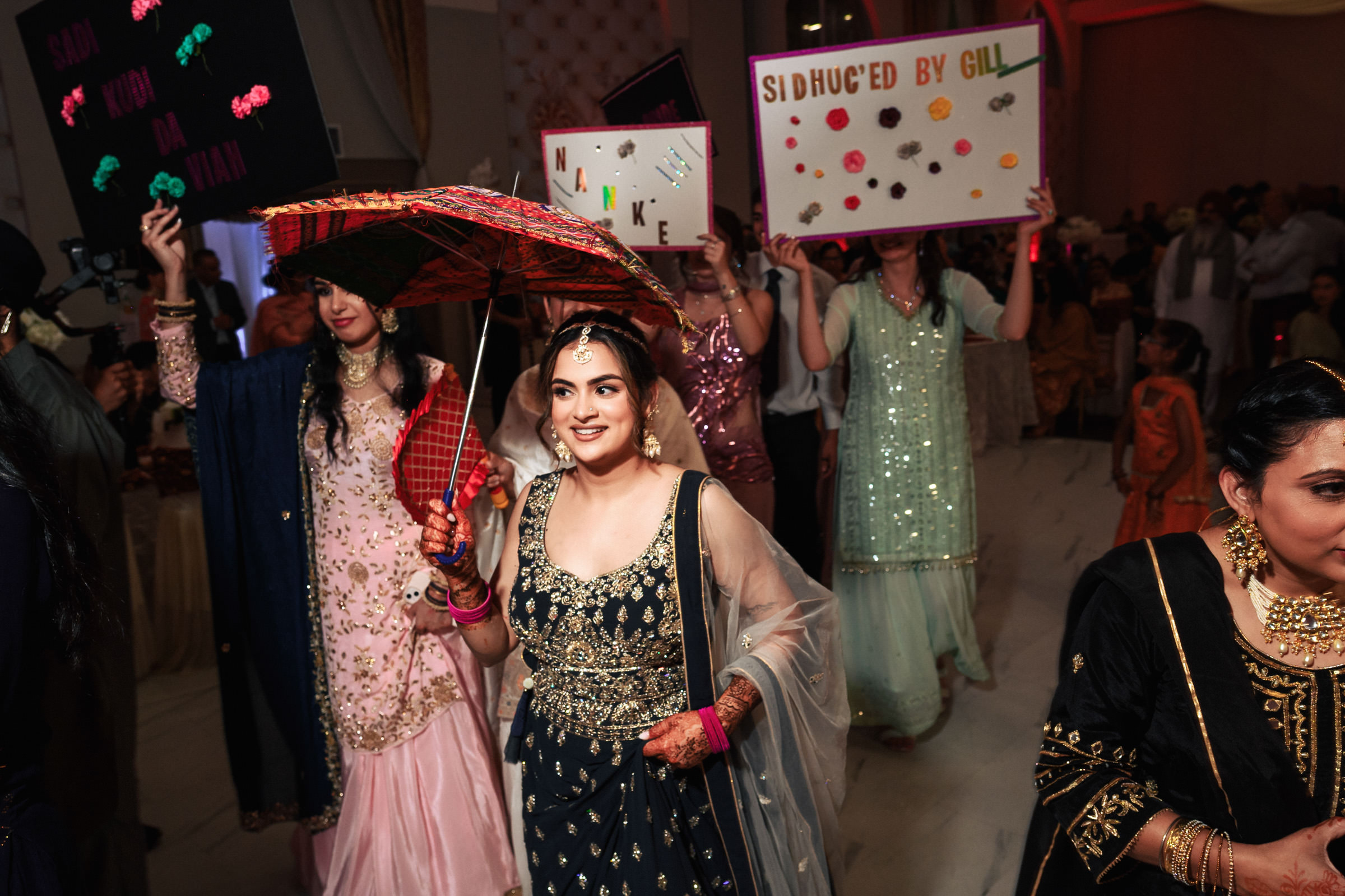 A woman in traditional attire smiles while holding a decorated umbrella. Behind her, people hold up signs and wear festive clothing in a lively, indoor celebration.
