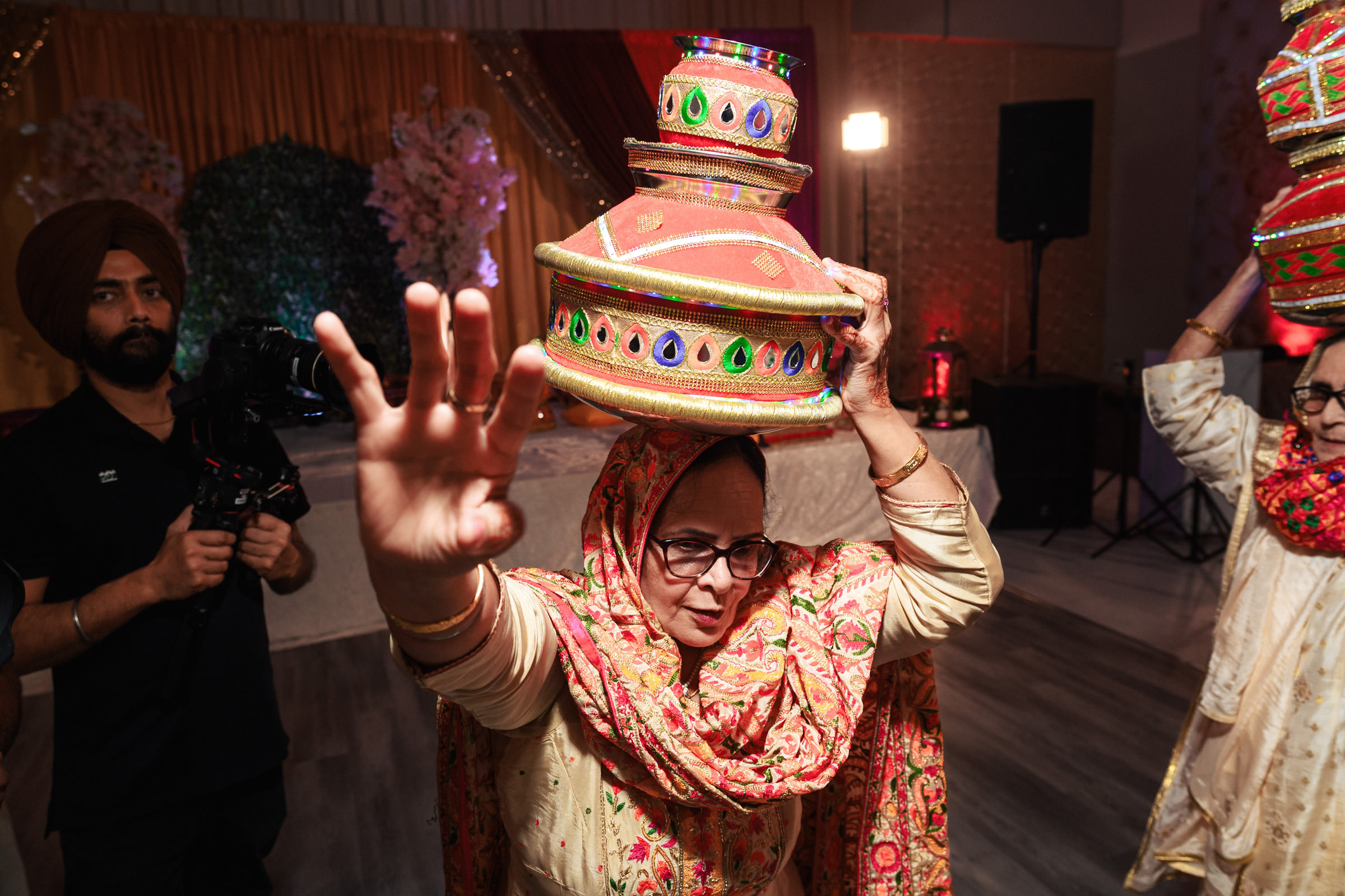 A woman wearing a traditional outfit and headscarf balances colorful, decorated pots on her head while dancing at an event. She is in mid-motion, with her hand extended forward. Another person dances nearby, and a photographer captures the scene.
