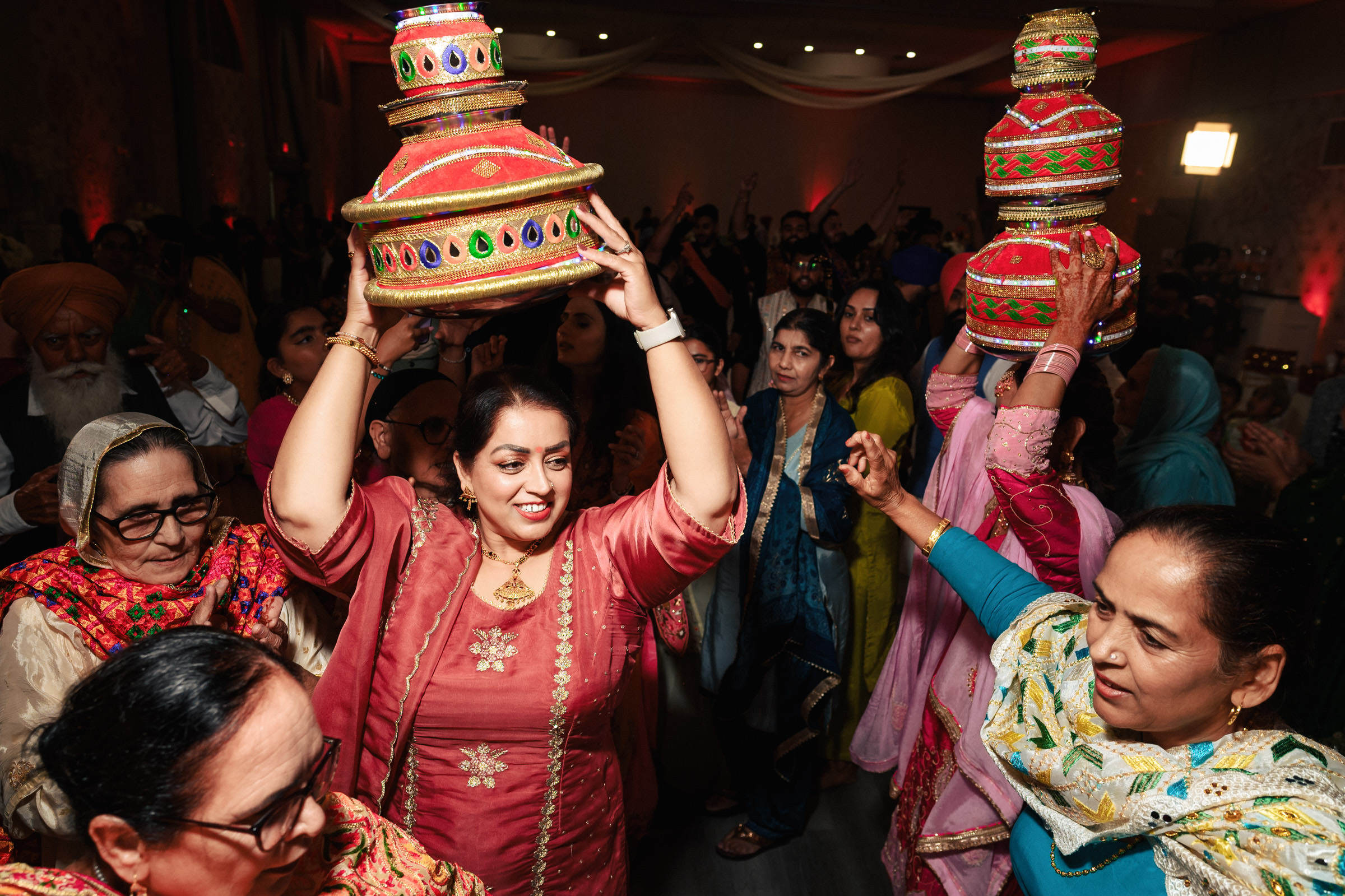 A group of women in colorful traditional attire dance and celebrate at an indoor event. They are holding vibrant, decorated vessels on their heads as part of the festivities. The atmosphere is joyful and lively.