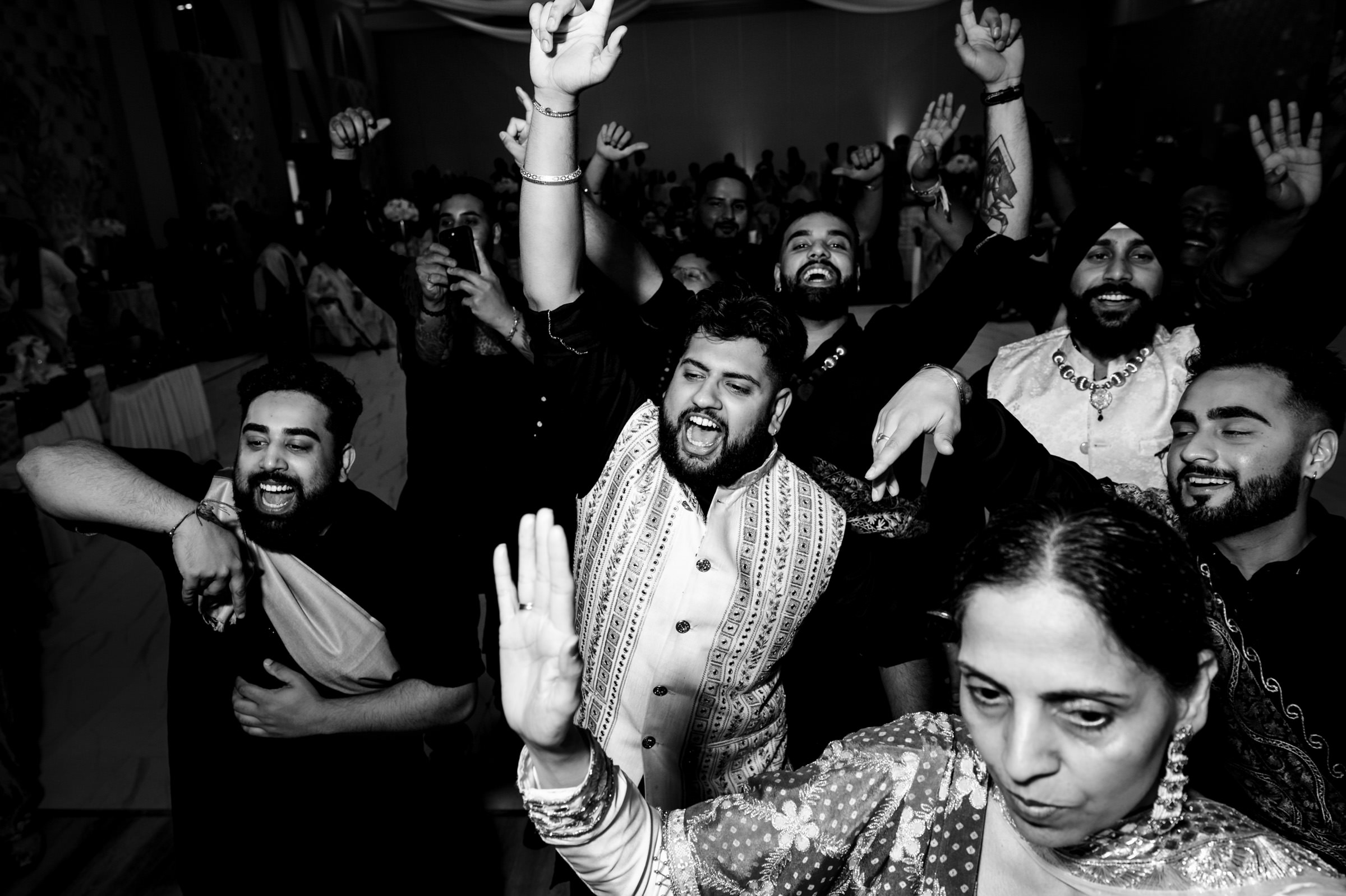 A lively group of people dancing energetically at a celebration, with joyful expressions and raised arms. The black and white photo captures the excitement and movement of the moment in a crowded indoor setting.