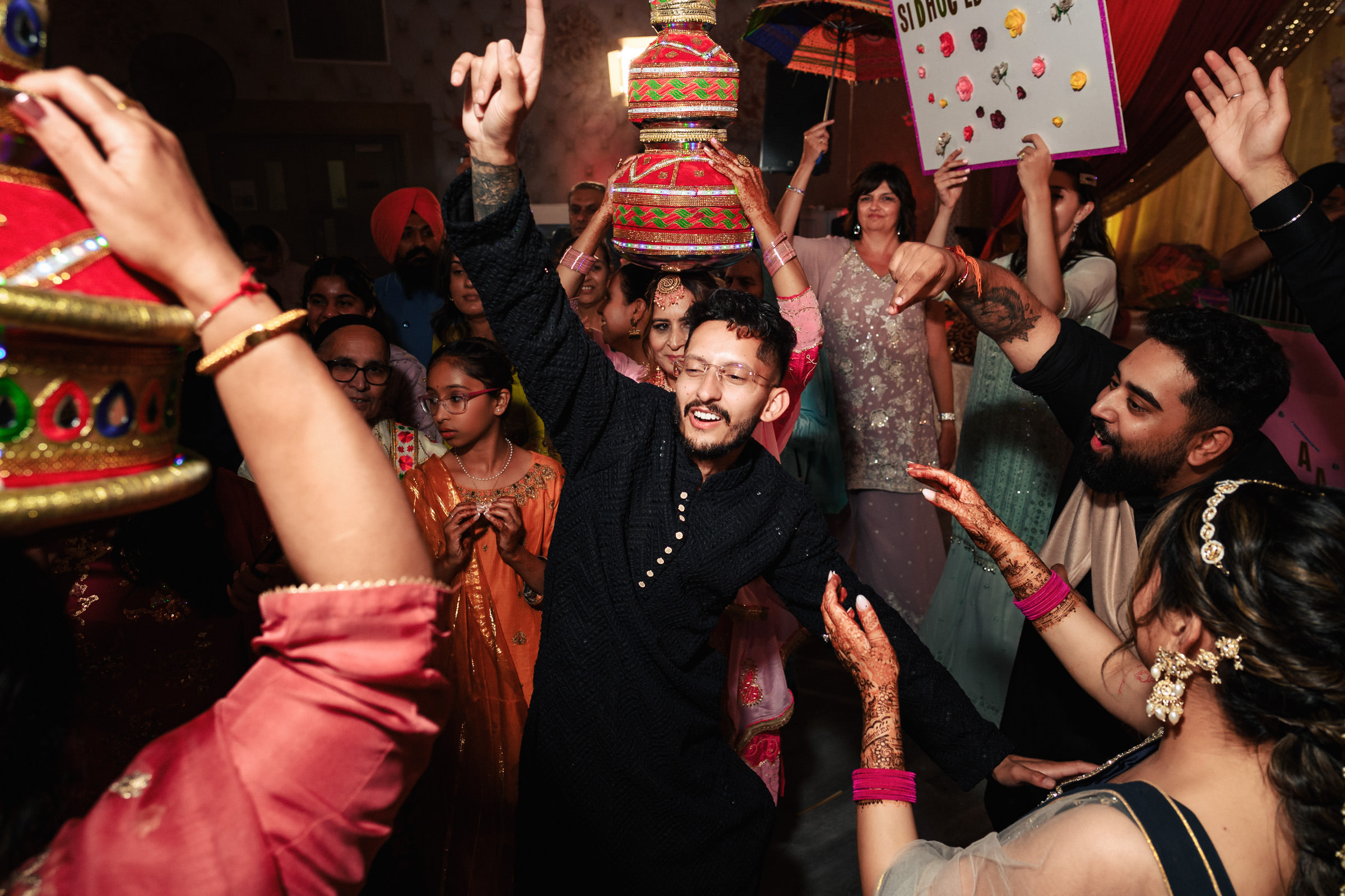 A joyful wedding celebration with people dancing energetically. The man in the center wears a black traditional outfit, surrounded by others in colorful attire, holding decorated pots and signs. Festive atmosphere with vibrant lighting.