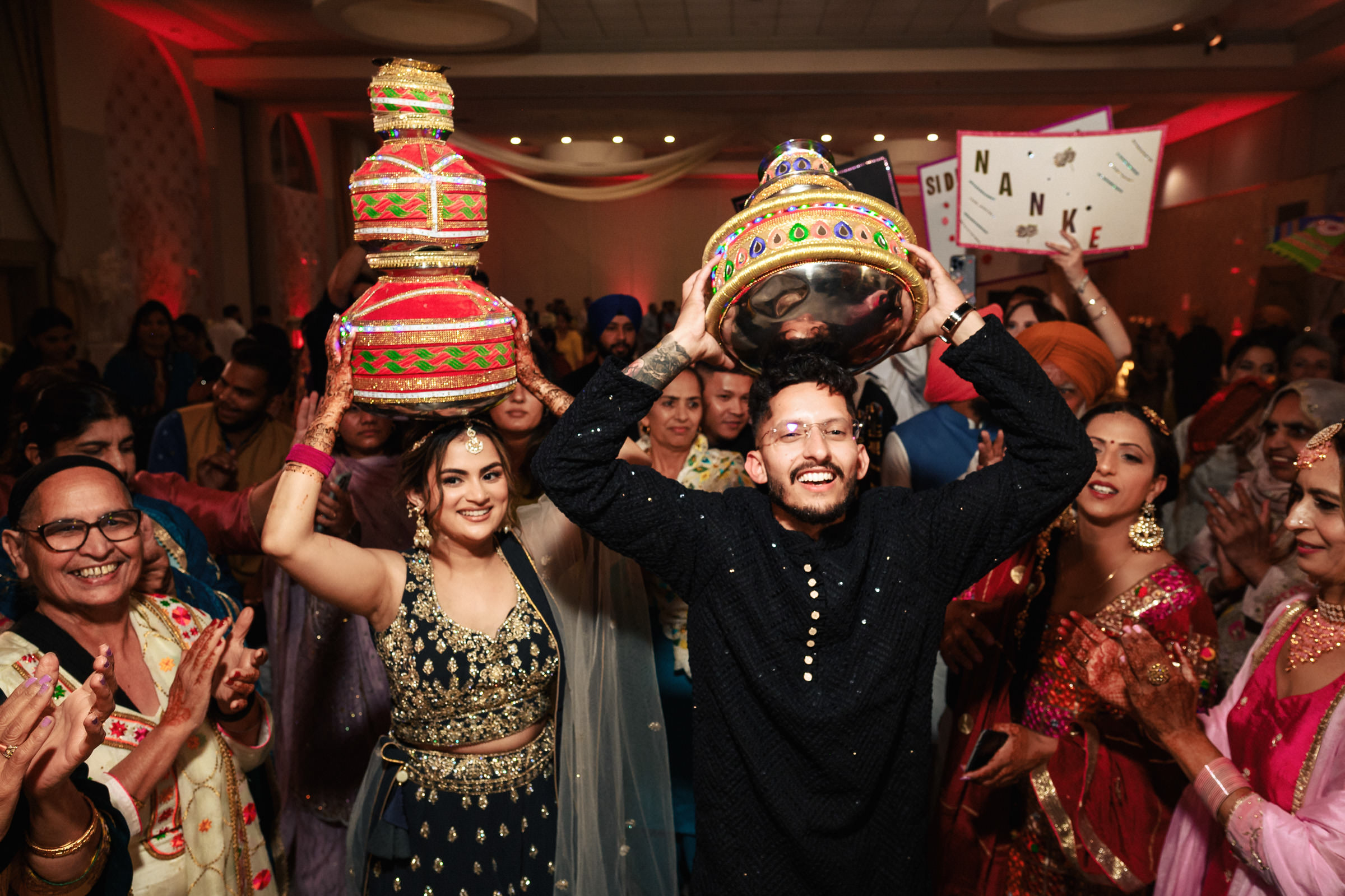 A group of people joyfully celebrates at an event. A man in traditional attire holds a colorful pot on his head, surrounded by women in festive clothing also with pots, dancing and smiling amidst a lively atmosphere.