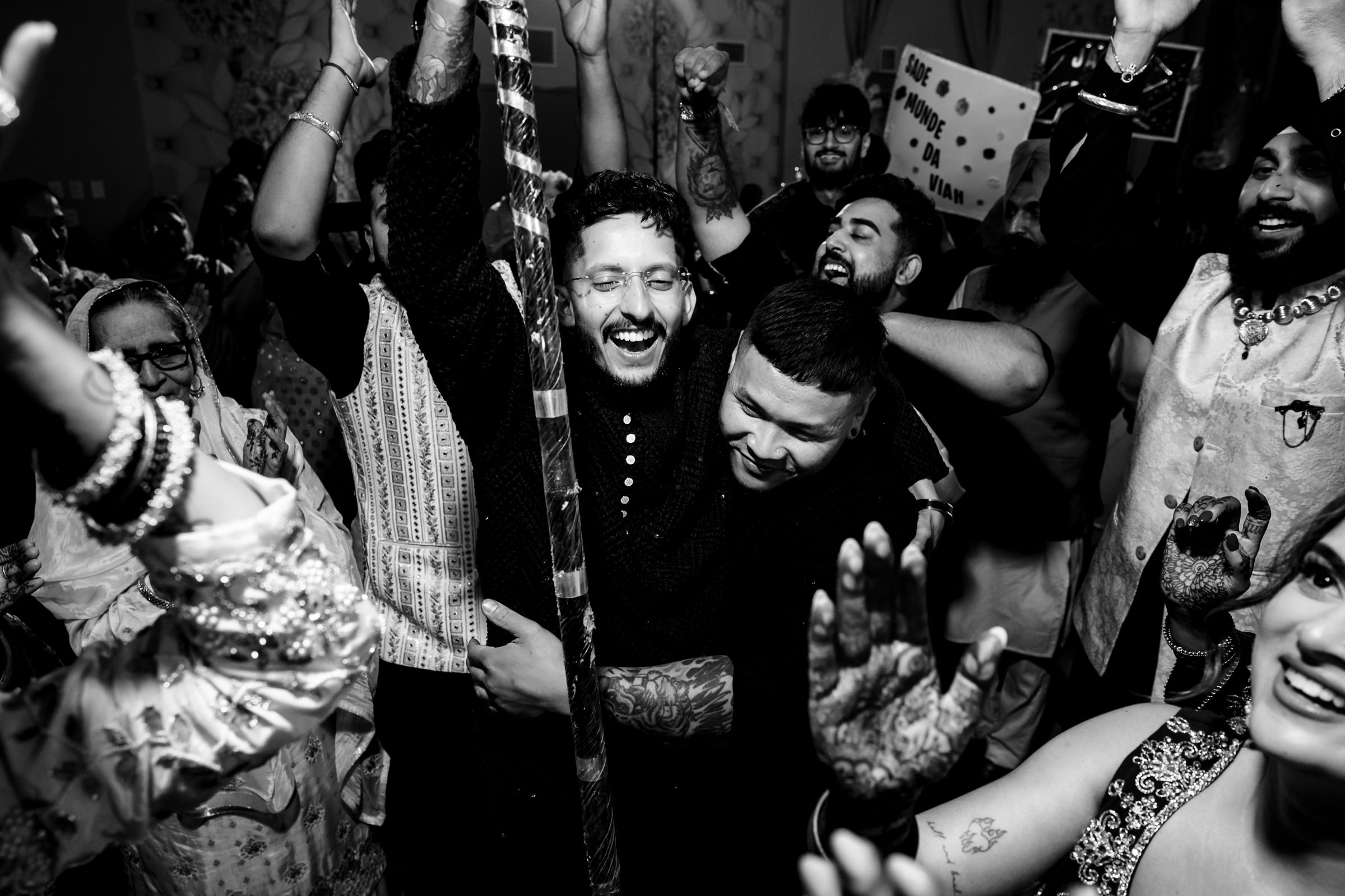 A joyful black and white scene of men and women celebrating at a party. Some are smiling and holding up their hands, while others embrace warmly. Traditional outfits and decorations are visible, adding to the festive atmosphere.