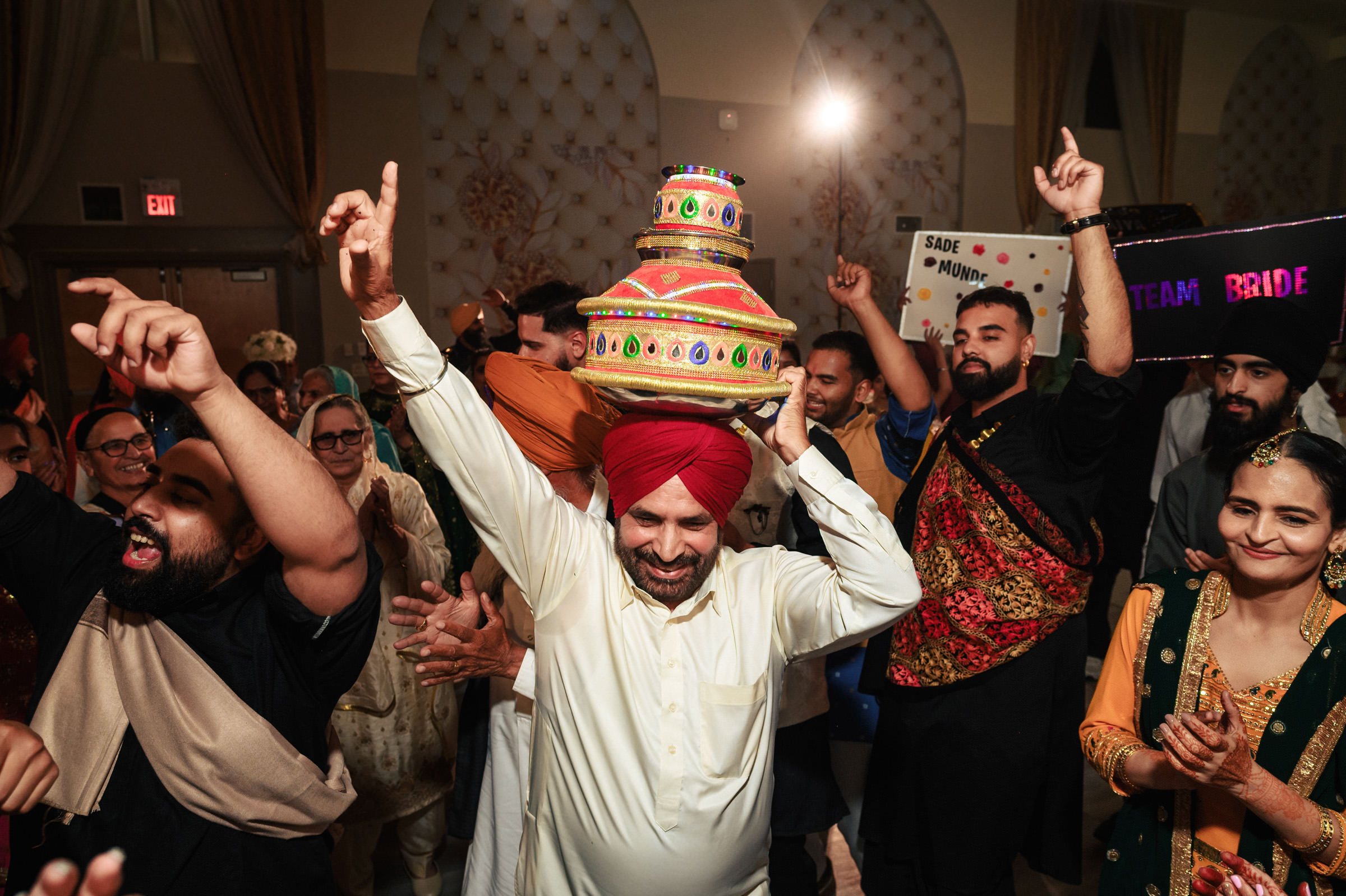 A joyous group of people is dancing at a festive event. A man wearing a red turban balances a colorful, decorated pot on his head. Others around him are smiling, clapping, and raising their hands in celebration.