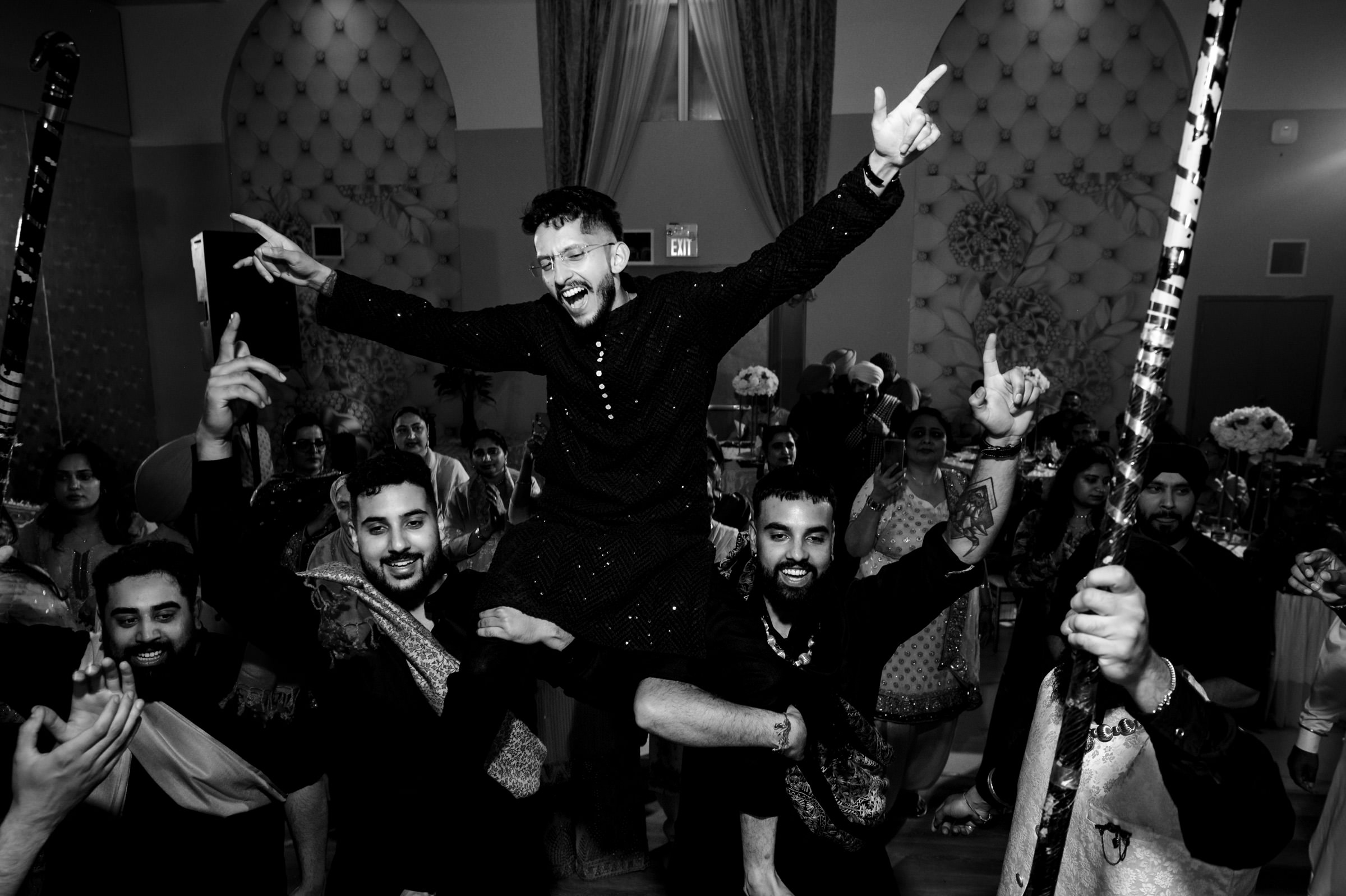 A joyous group of people celebrates indoors. A man is lifted on shoulders, smiling widely, with arms raised. Others around him hold decorated sticks, wearing traditional clothing, in a lively dance atmosphere. Black and white photo.