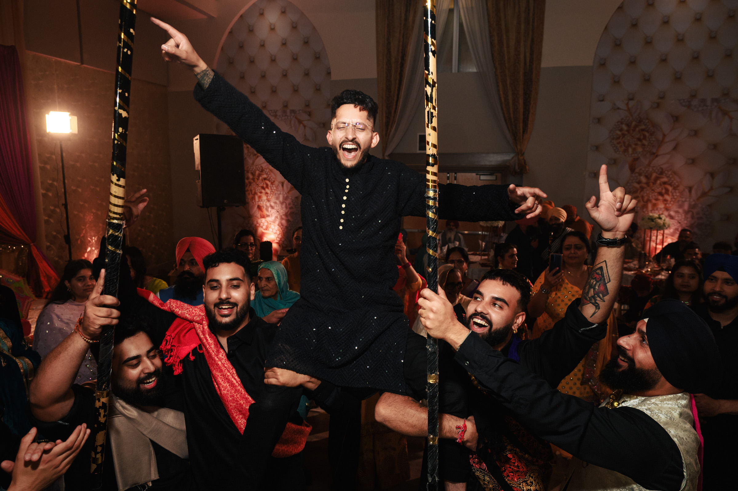 A group of men in traditional attire lift a joyful man onto their shoulders at a vibrant celebration. The background is filled with people watching and ornate decorations, creating a festive atmosphere.