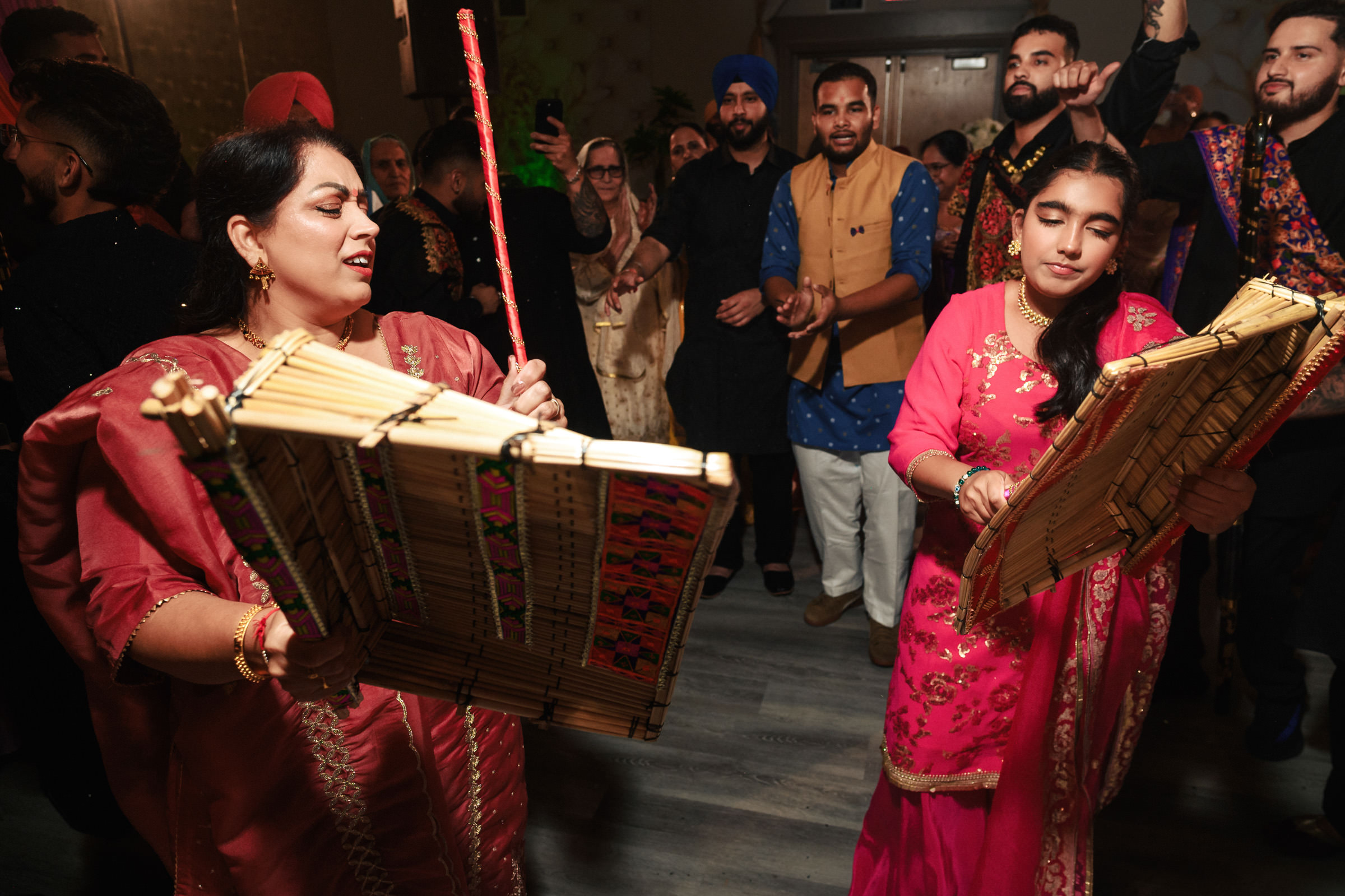 Two women in traditional Indian attire dance with wooden instruments at a lively gathering. They are surrounded by people clapping and watching, dressed in colorful festive clothing. The atmosphere is joyful and vibrant.
