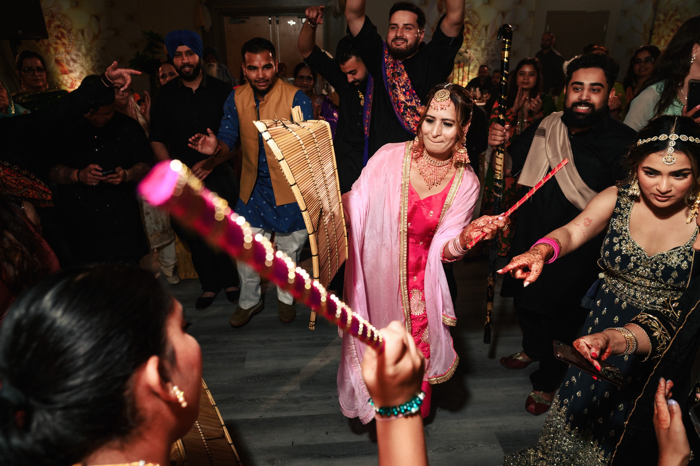 A vibrant celebration with people dancing energetically, holding colorful sticks. A woman in a pink outfit leads the group, surrounded by others in festive attire. The atmosphere is joyful and lively, with intricate costumes and decor enhancing the scene.