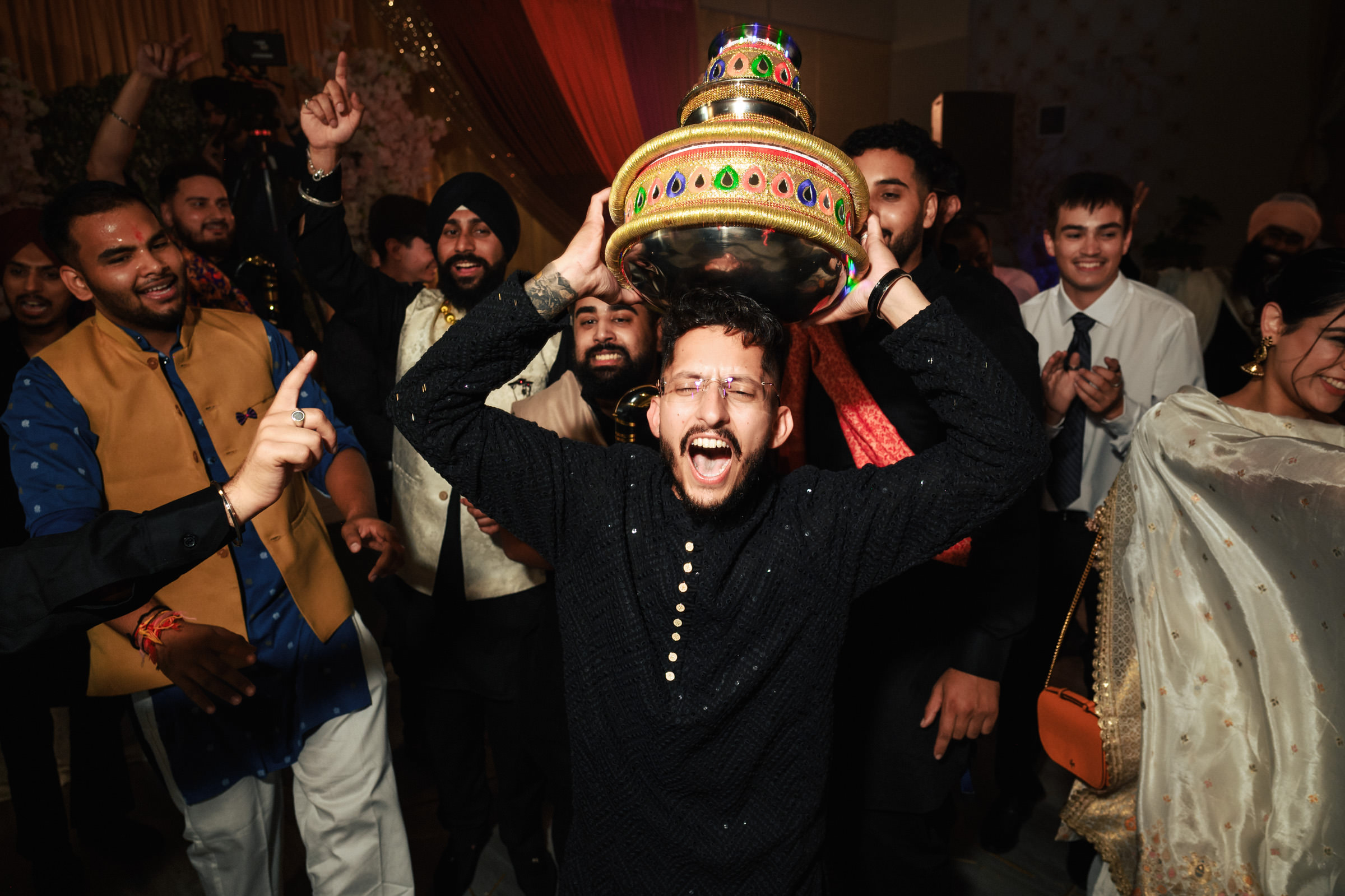 A joyful man, wearing glasses and a black sweater, holds a decorated pot on his head while smiling widely. He is surrounded by a group of people celebrating, with some clapping and cheering, in a festive, warmly lit setting.