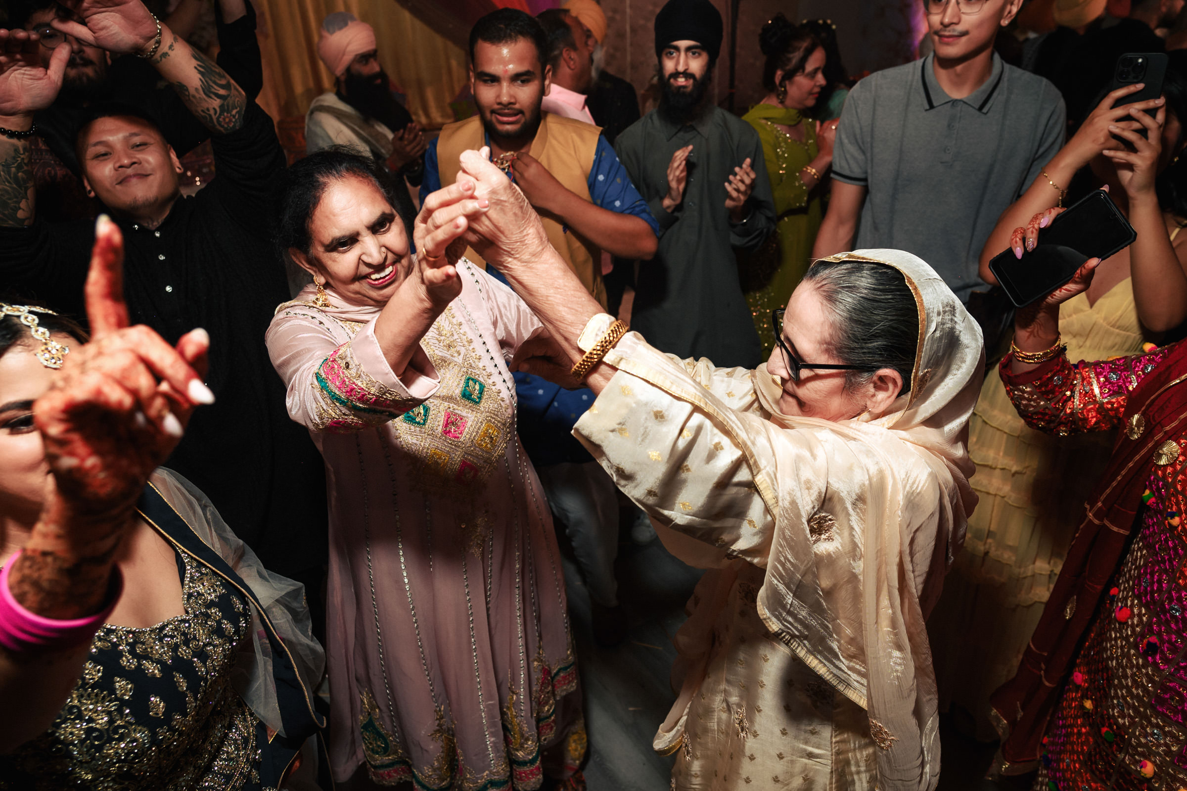 A lively group of people celebrating and dancing at an event. An elderly woman in traditional attire is dancing joyfully with another woman. Others around them are smiling, clapping, and capturing the moment on their phones.