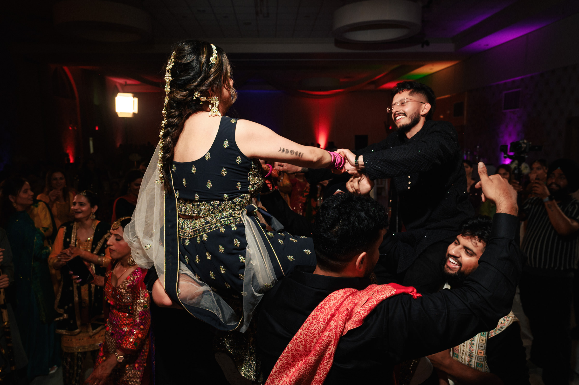 A lively celebration with people dancing in traditional attire. A man and woman, elevated by others, hold hands and smile at each other. The room is dimly lit with colorful lighting, and the group is surrounded by cheering onlookers.