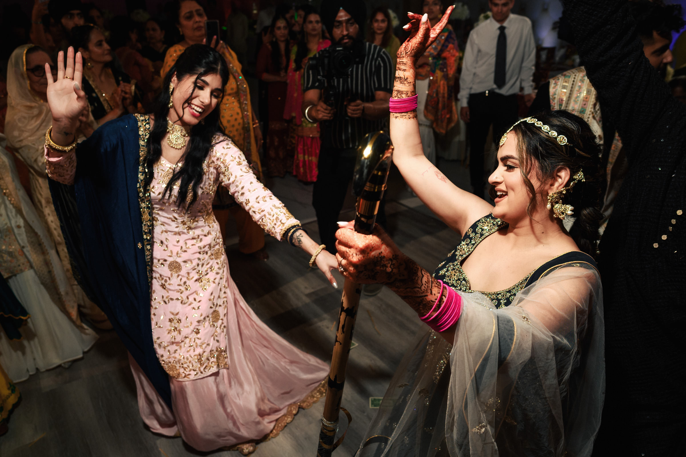 Two women joyfully dance at a lively celebration. One wears a pink outfit and the other a green one, both adorned with intricate henna. They're surrounded by a crowd of people, creating a festive atmosphere. Both hold sticks, possibly for a traditional dance.