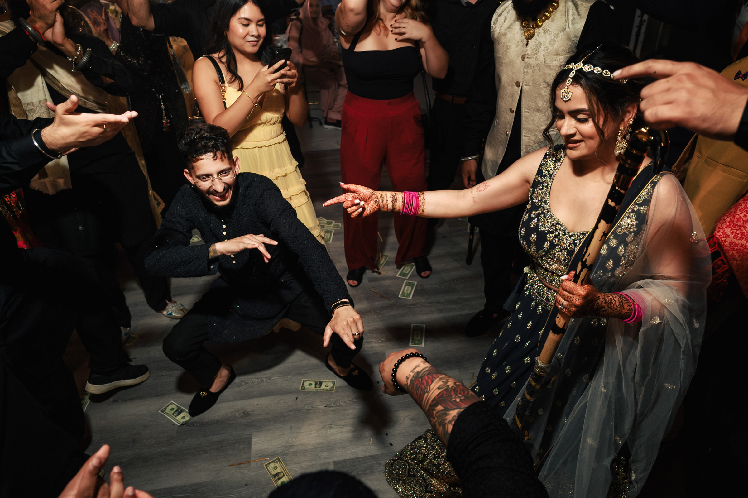 A lively dance floor scene with a man crouching and smiling at a woman dressed in ornate traditional attire holding a staff. Other people clap and watch as dollar bills lie on the floor. The atmosphere is festive and energetic.