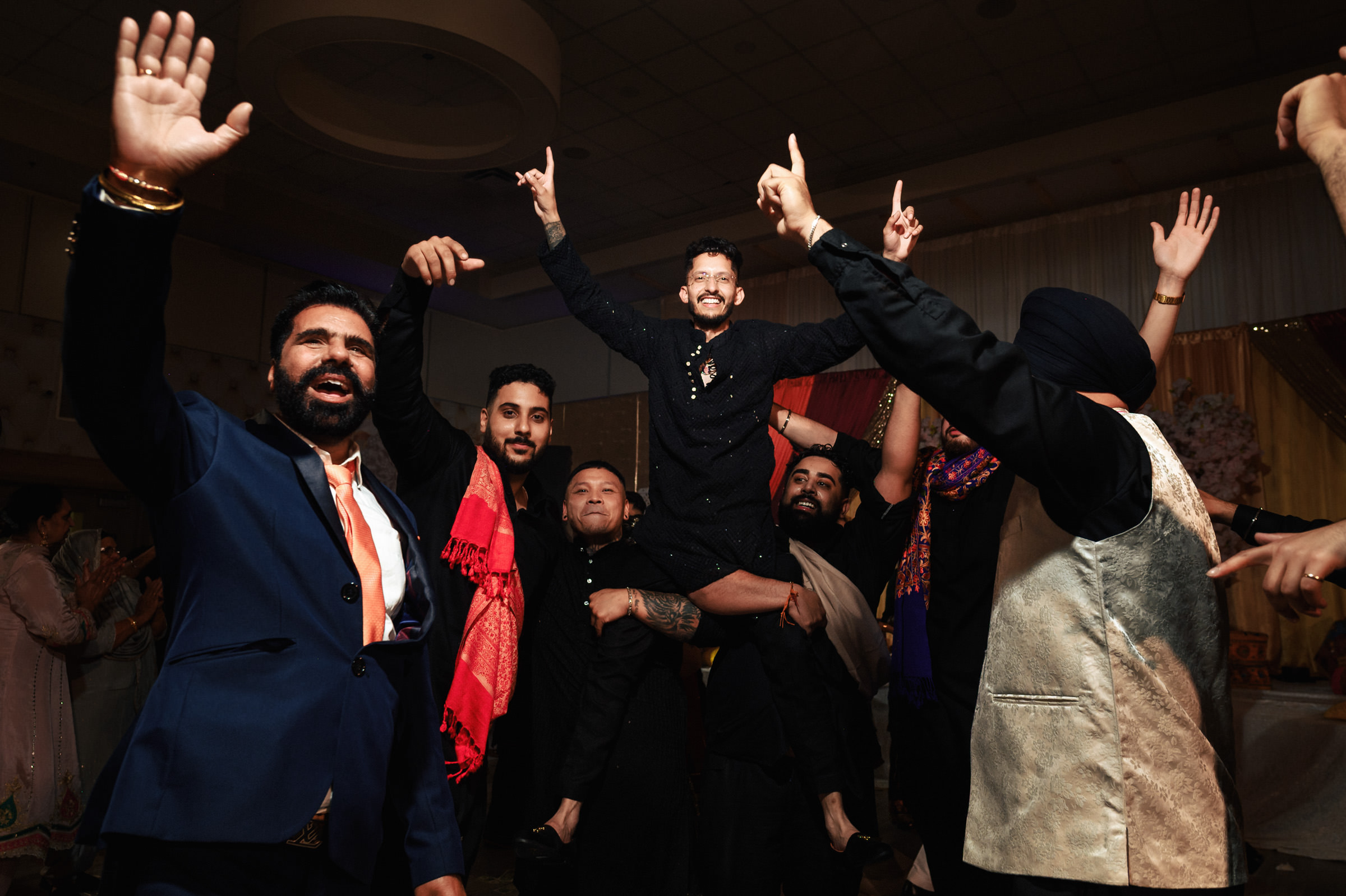 A group of people celebrates at an indoor event. A man is lifted in the air by others while smiling and raising his hands. The atmosphere is joyful, with attendees wearing formal attire and vibrant scarves.