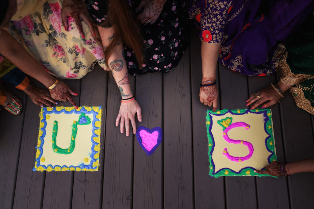 Five people display painted banners on a dark wooden floor. Two banners, marked with "U" and "S" in colorful designs, flank a central heart symbol. The participants wear brightly colored traditional attire and display intricate henna patterns on their hands.