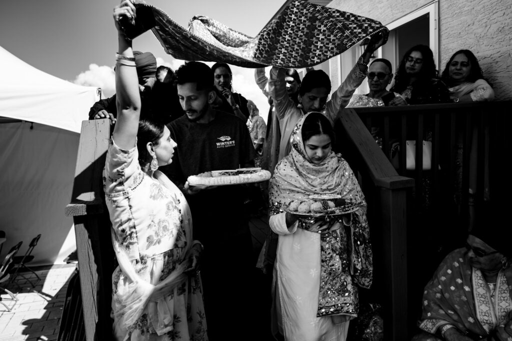 A black and white photo of a wedding ceremony. The bride, in traditional attire, is stepping down stairs with a decorative cloth held over her. A woman holds a tray beside her, surrounded by a small group of people, some in traditional attire.